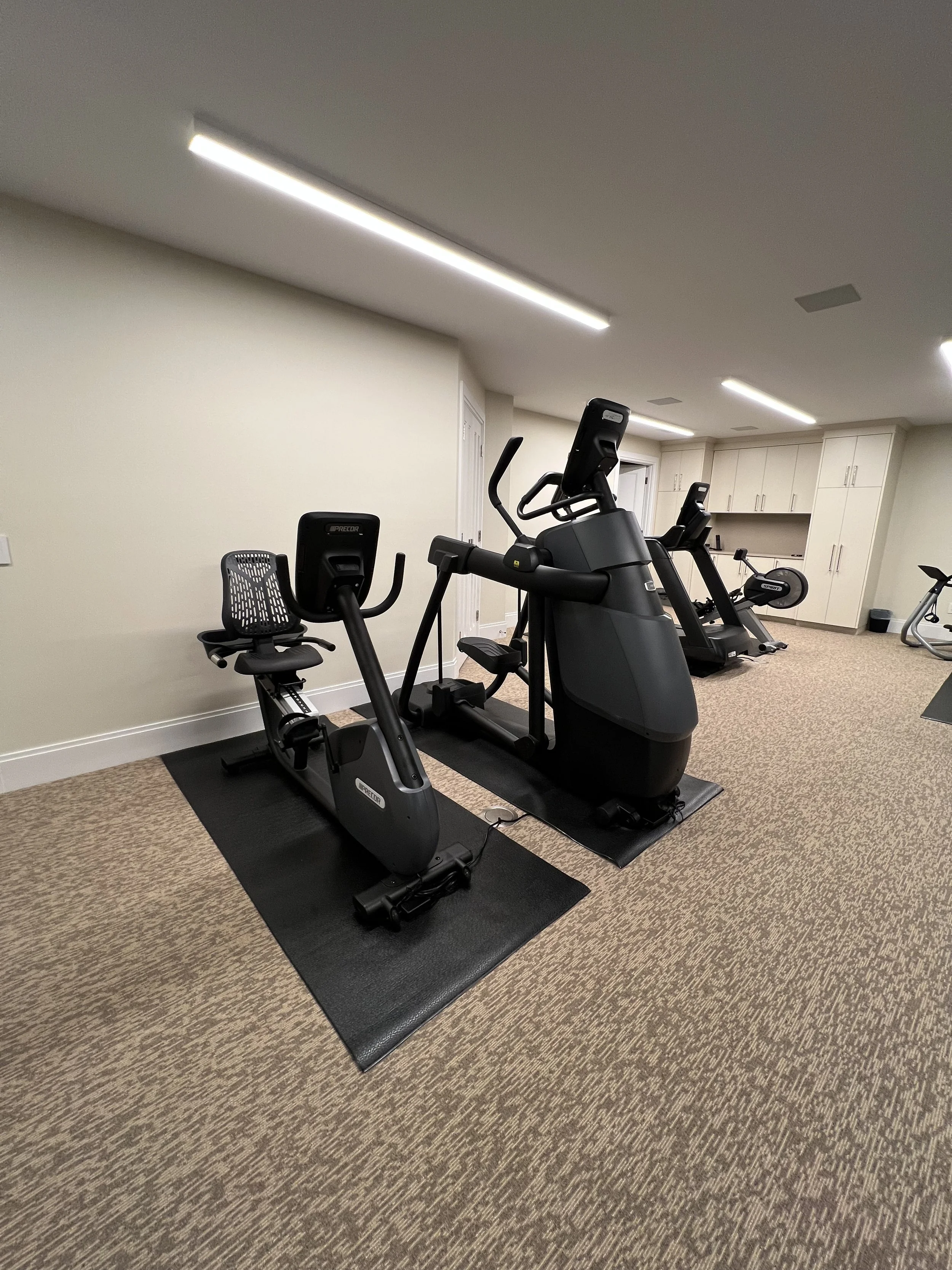 A home gym with a recumbent bike, an elliptical trainer, and a treadmill on a tan carpeted floor with ceiling lights and white cabinets in the background.