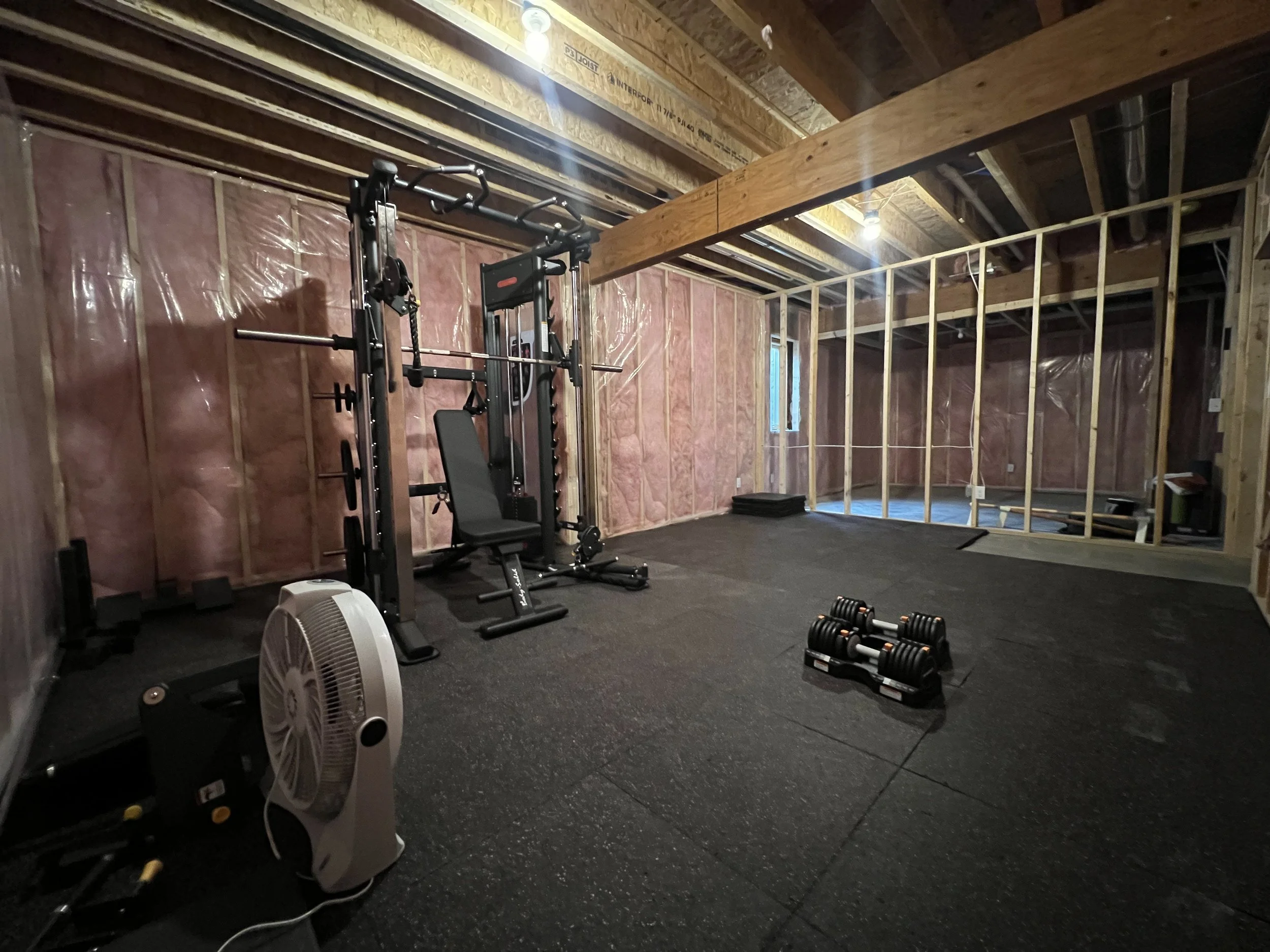 Empty home gym with a weight machine, a set of dumbbells, a standing fan, and a small window in a partially unfinished room with pink insulation on the walls and exposed wooden beams on the ceiling.