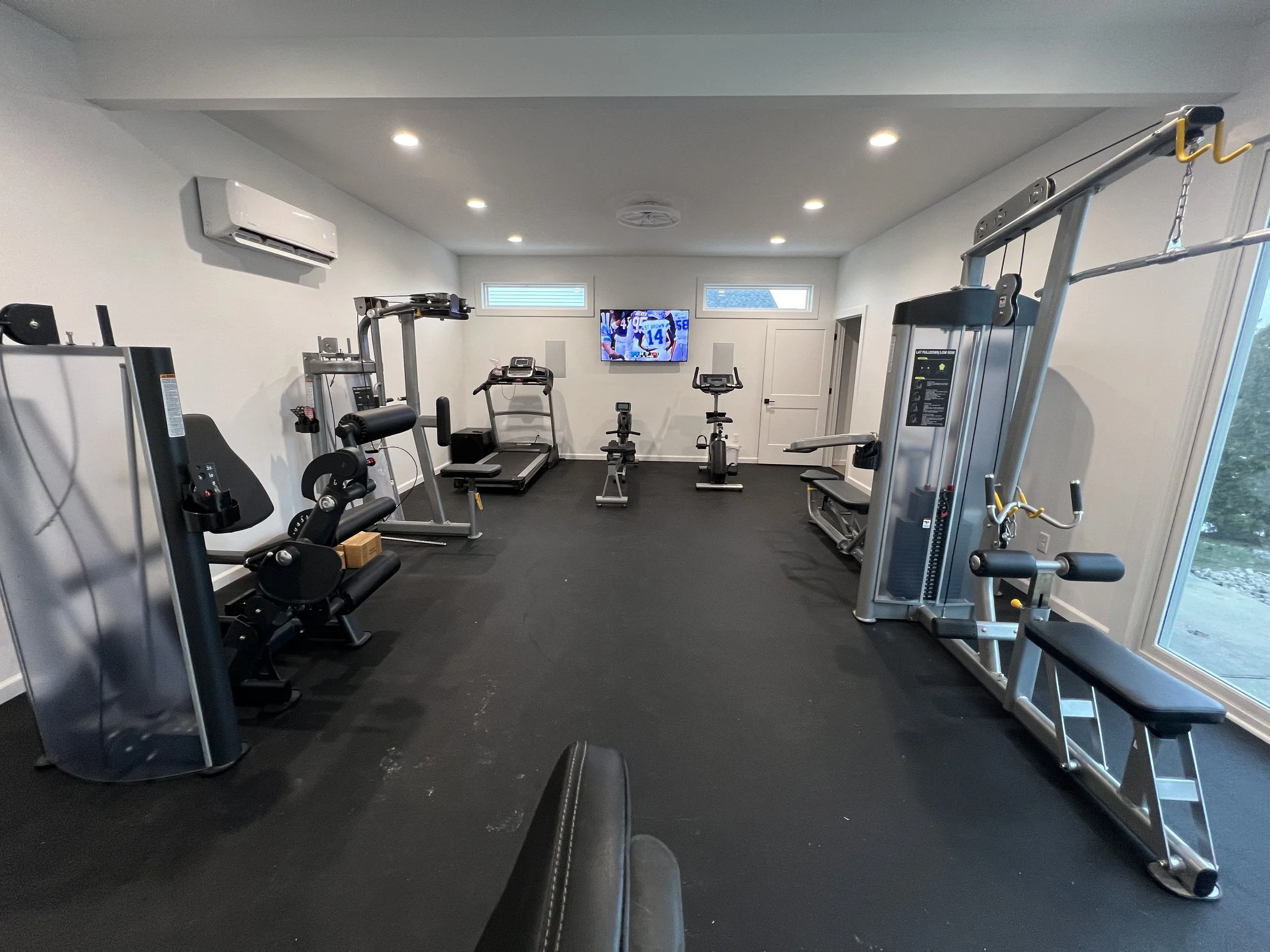 Empty home gym with exercise equipment including treadmill, stationary bike, weight machine, and bench, with a wall-mounted TV and windows.