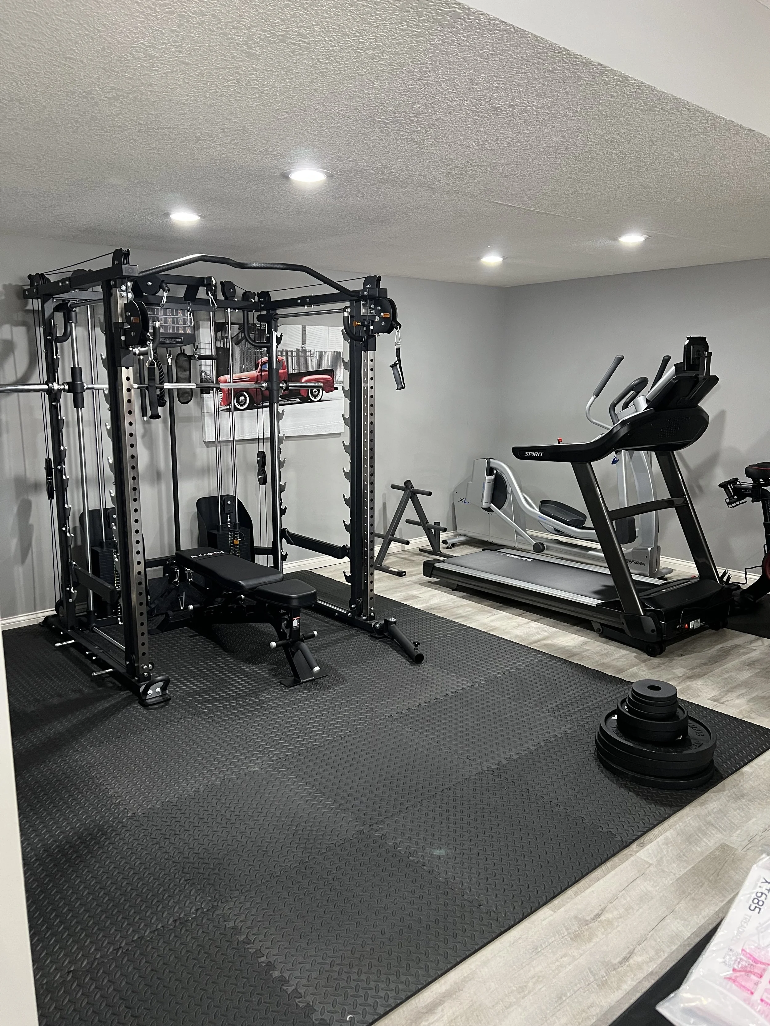 Home gym with a black multi-exercise machine, a treadmill, a stationary bike, a small exercise bench, stacked weight plates, and black rubber flooring.