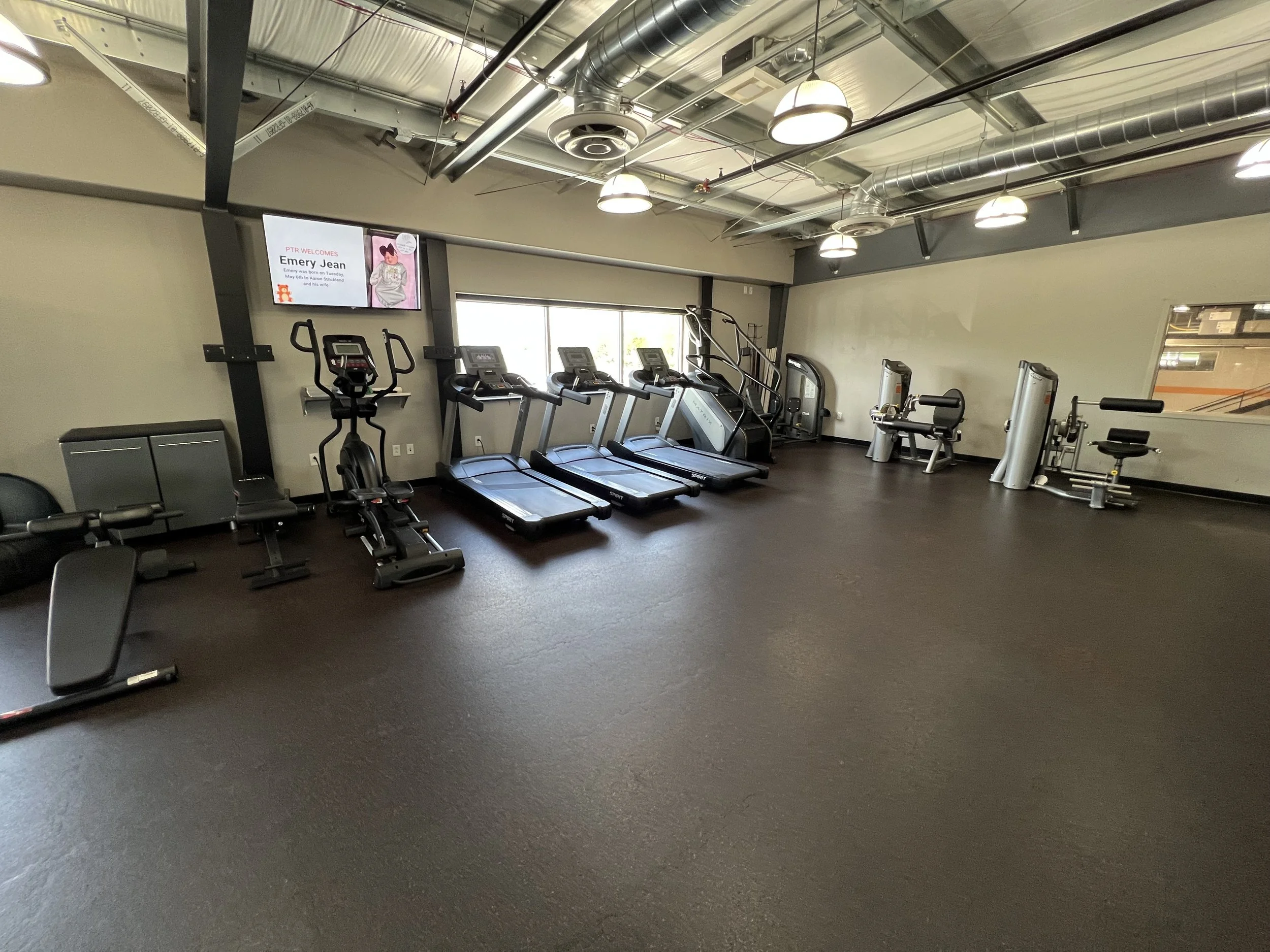 An empty gym with various exercise equipment, including treadmills, a stationary bike, weight machines, and a bench. There is a large window providing natural light and a wall-mounted display screen on the left.