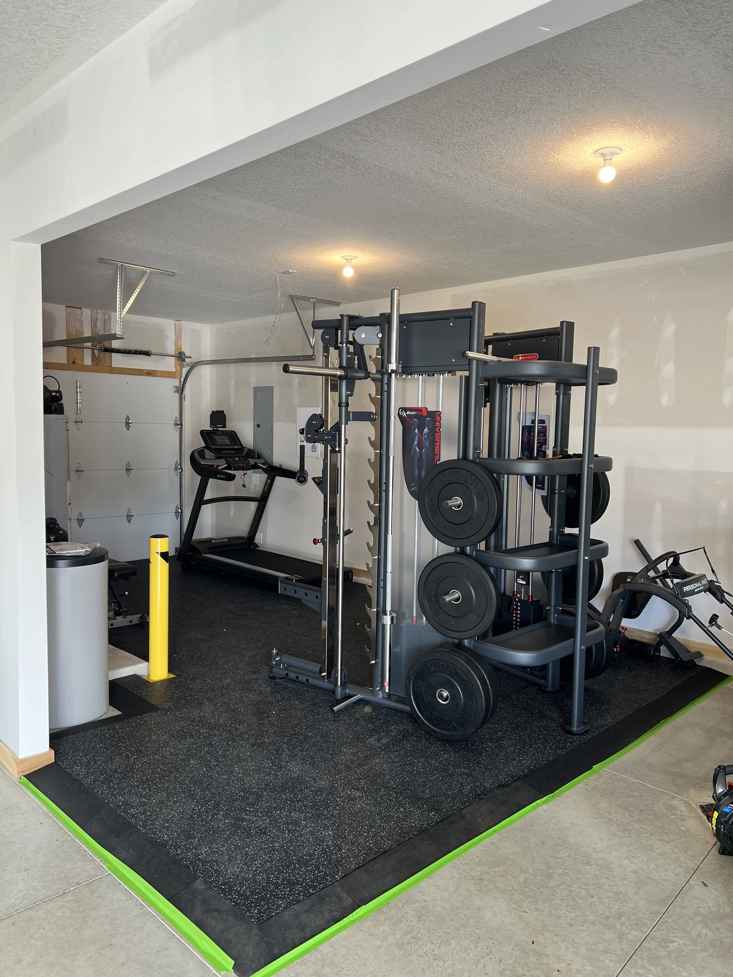 Home gym with treadmill, weight rack with weights, and other fitness equipment on black rubber flooring with green border.