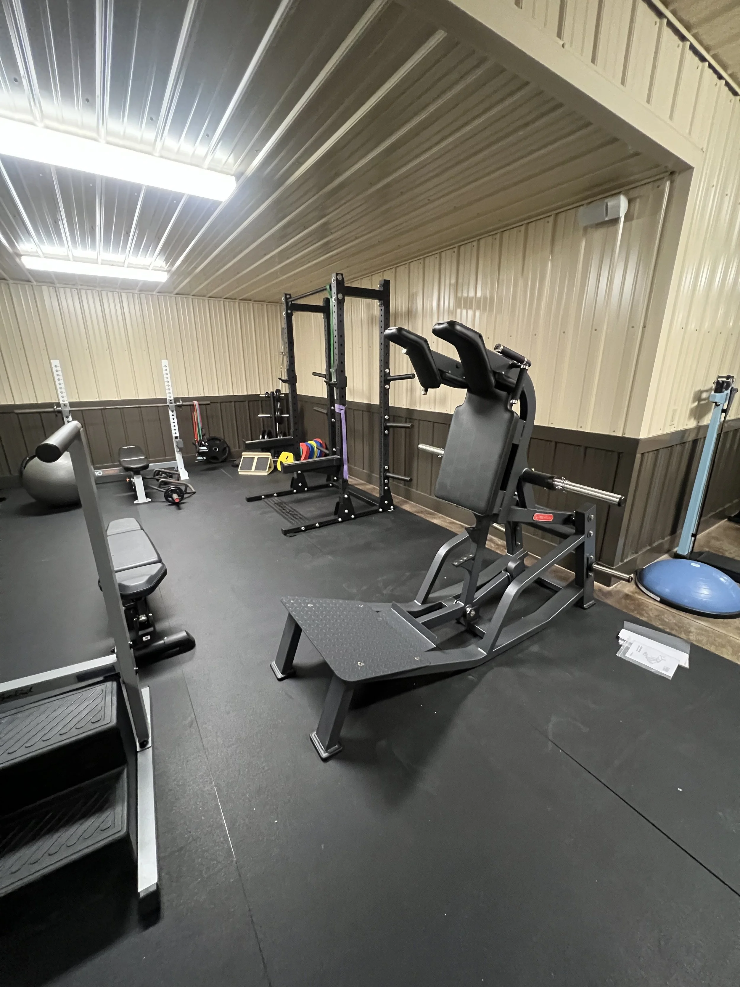 Home gym with various workout equipment, including a leg press, weight rack, weight plates, exercise ball, and a blue balance trainer within a small room with wooden paneled walls and ceiling lighting.