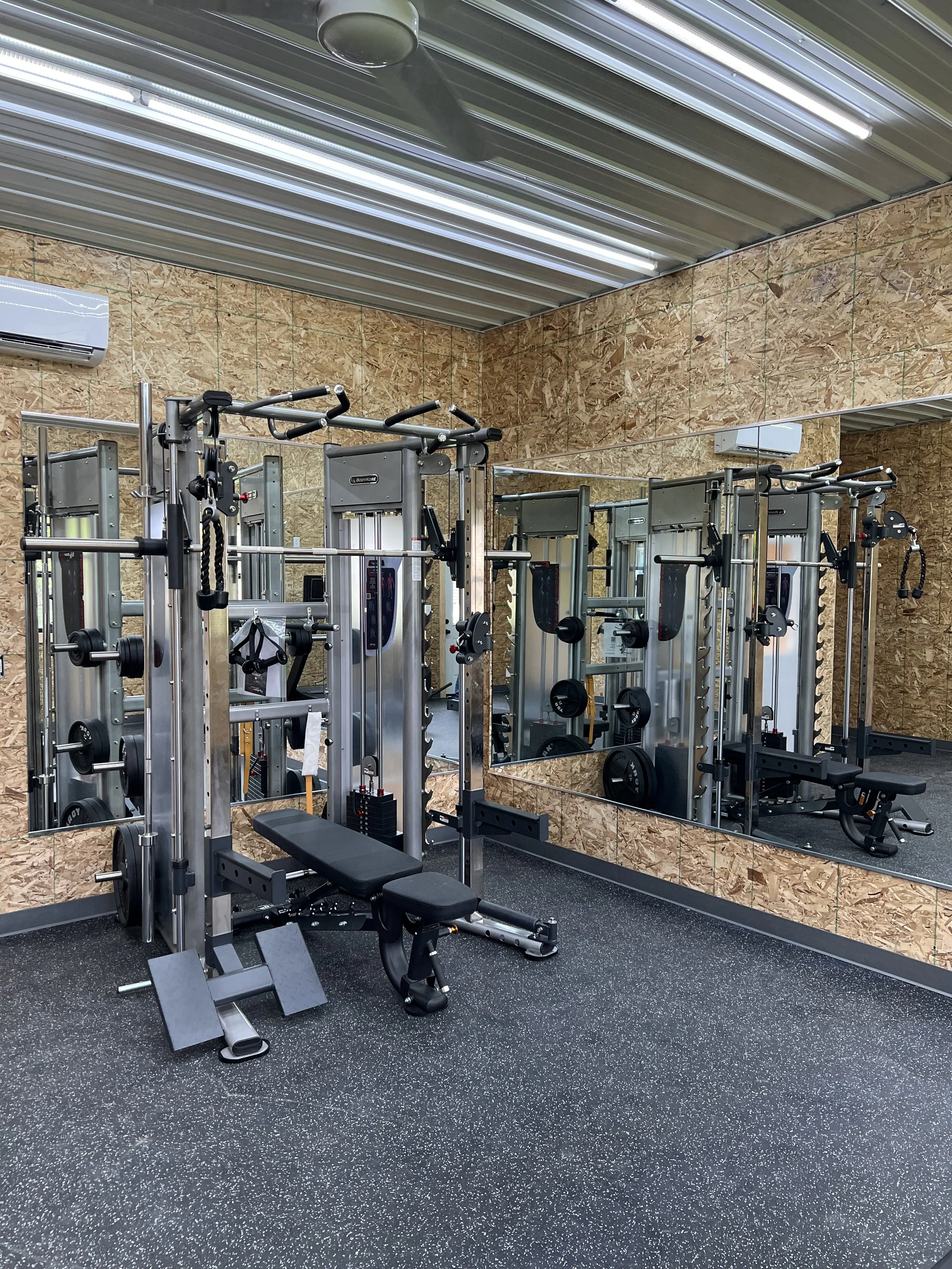 A small gym with workout equipment including weight machines, a flat bench, and mirrors on the wall, with wood paneling and a rubber flooring.