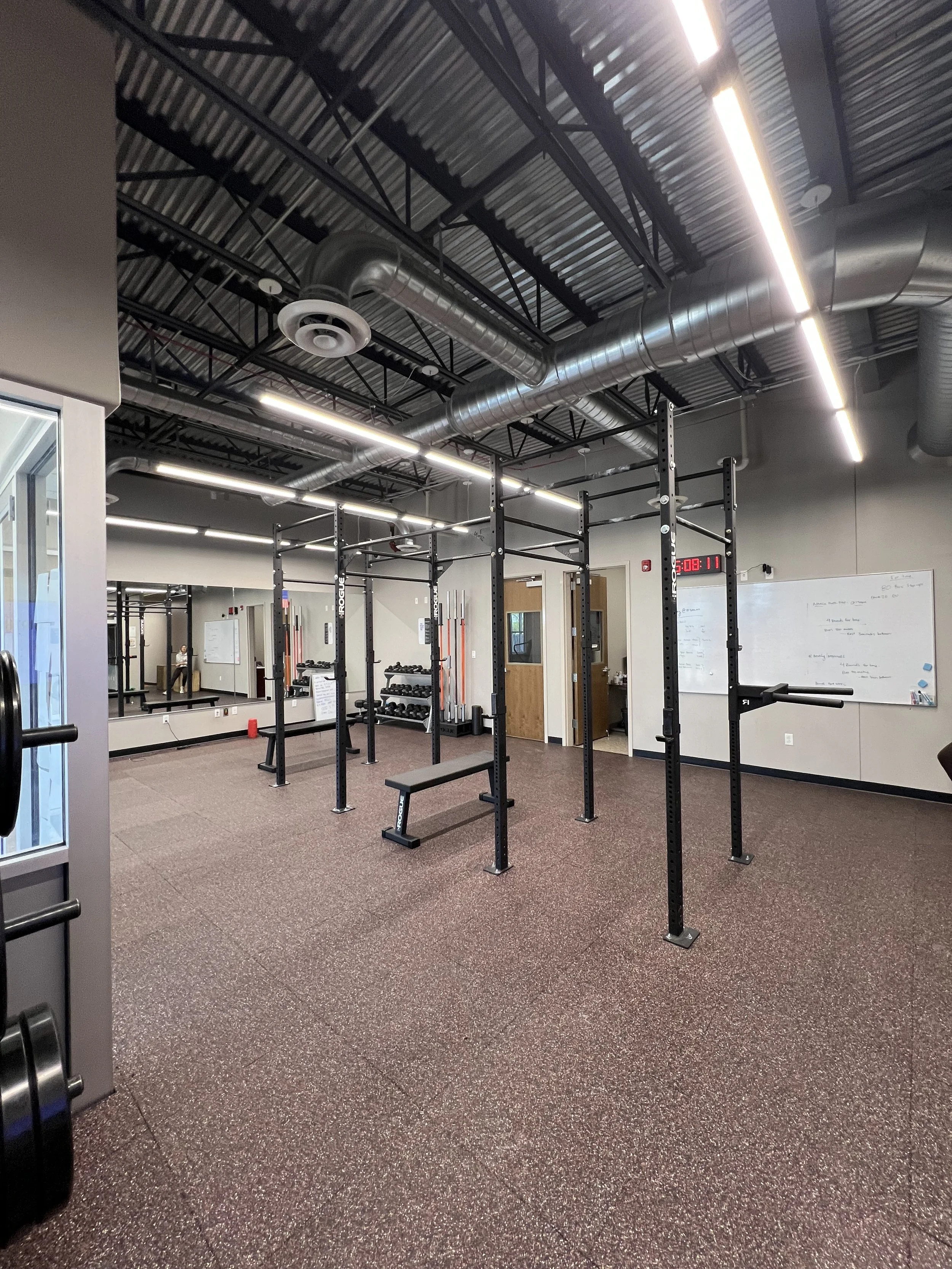 An empty gym with weightlifting and CrossFit equipment, including pull-up bars, a bench, dumbbell racks, and mirrored walls, with a digital clock displaying 5:08.