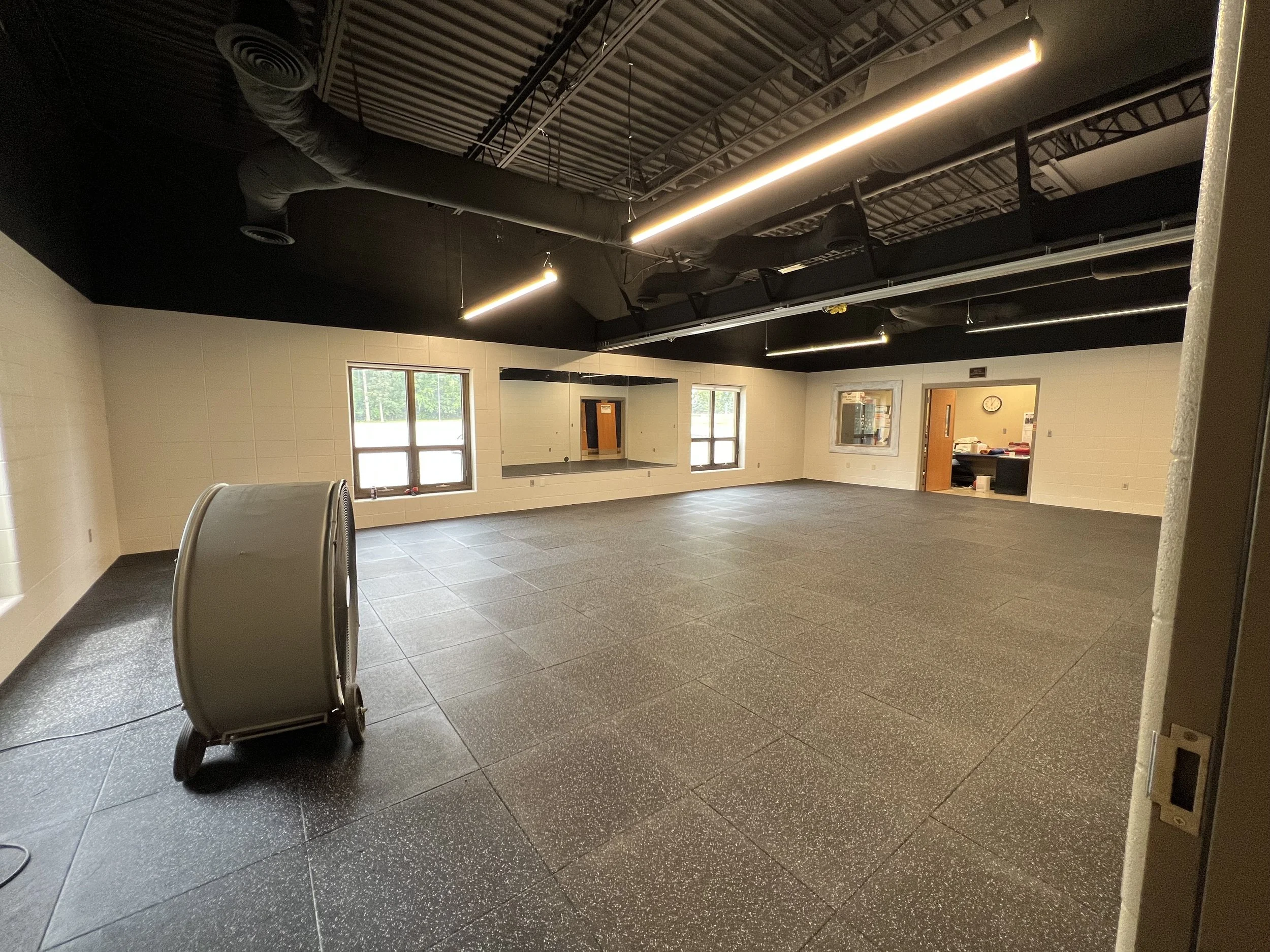 Empty gym or multipurpose room with black flooring, beige walls, large mirror, windows, and industrial ceiling with black ducts and lighting fixtures.