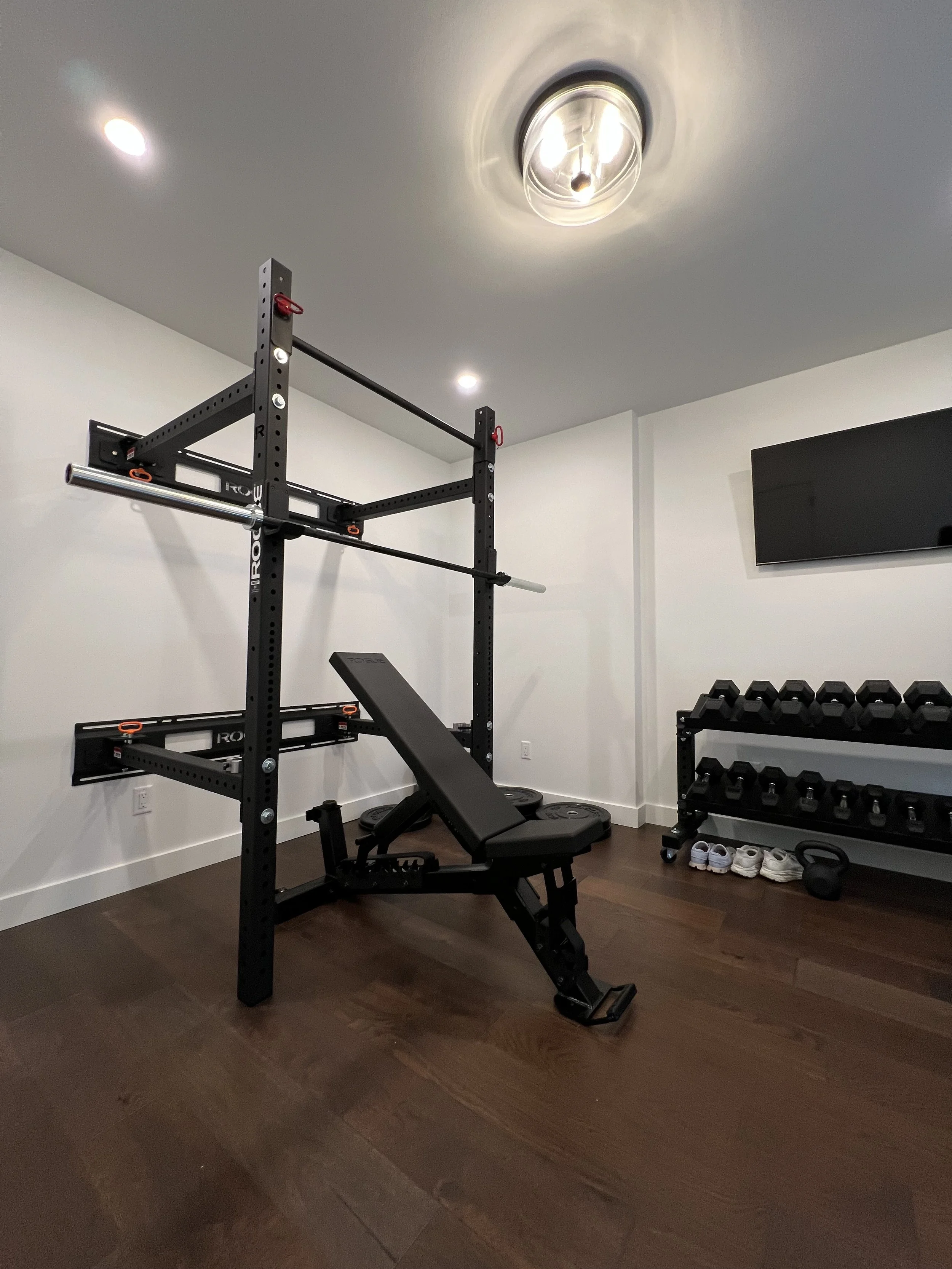 Home gym room with a power rack, adjustable weight bench, dumbbells, and a television on the wall.
