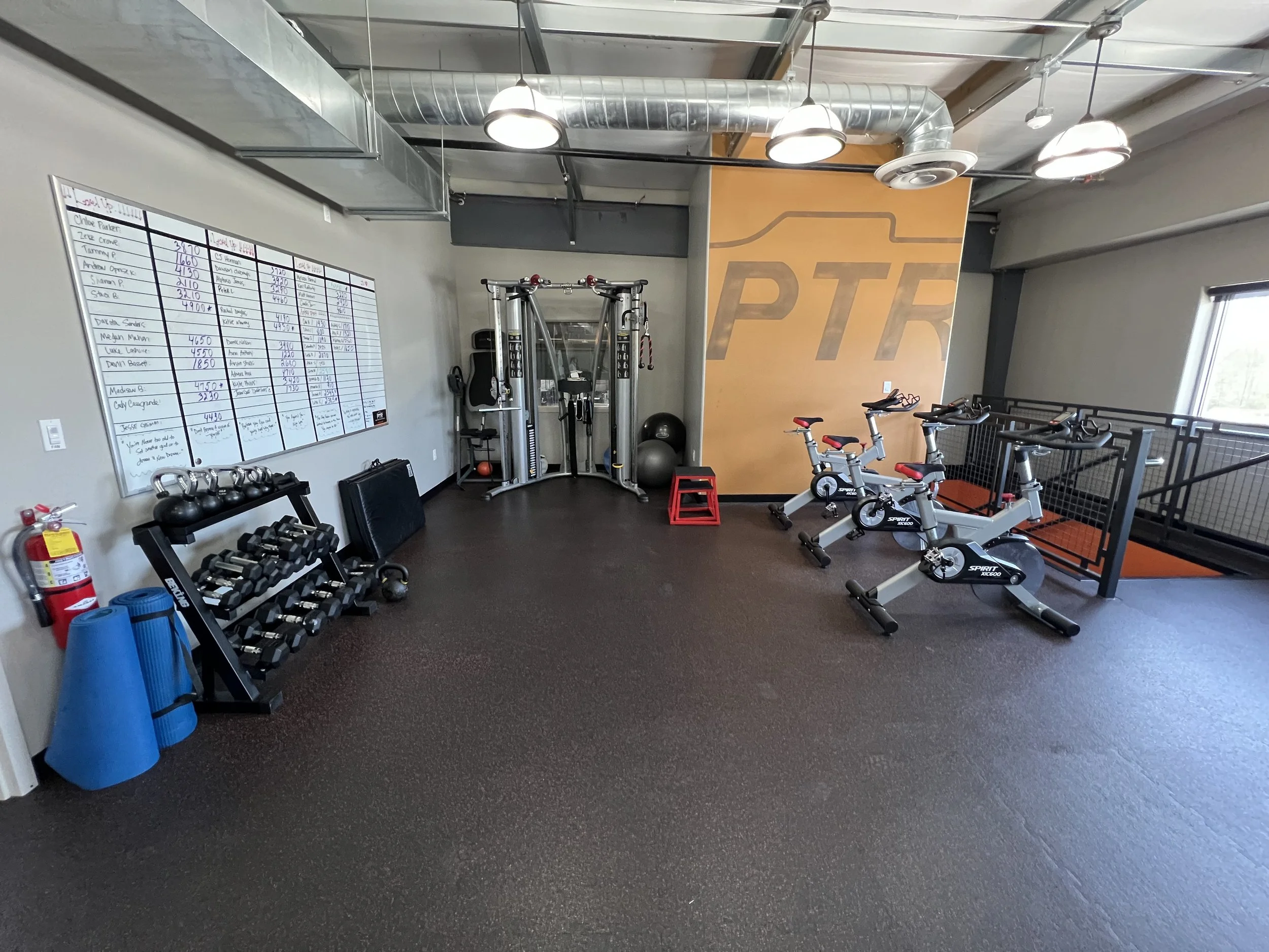 Fitness room with three exercise bikes, a weight rack, kettlebells, dumbbells, a whiteboard with workout routines, and exercise balls. The room has a black rubber floor, industrial ceiling, and a large logo on an orange wall.