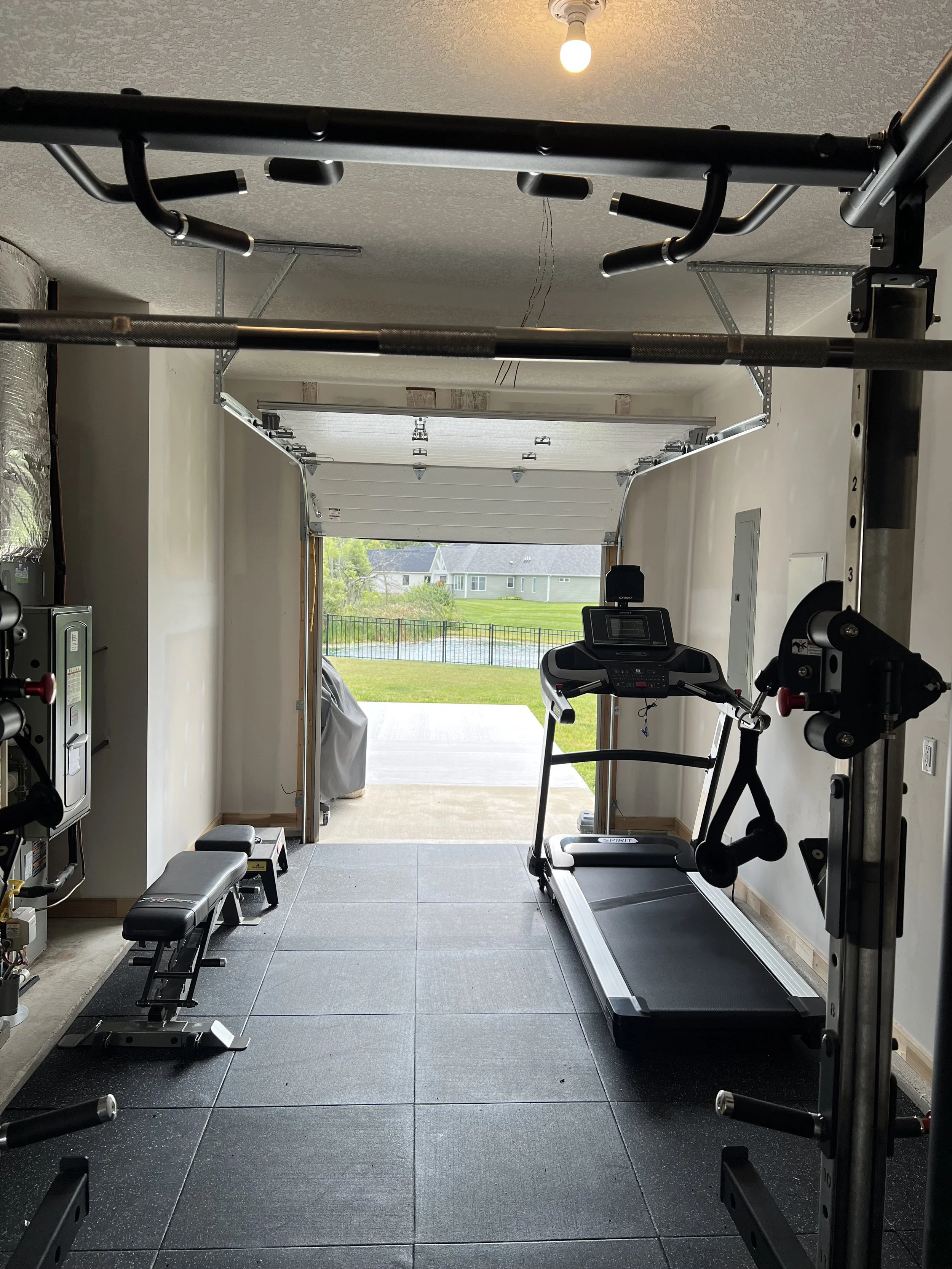 Home gym with treadmill, weight bench, and pull-up bar, overlooking a backyard with grass, fence, and houses in the distance.