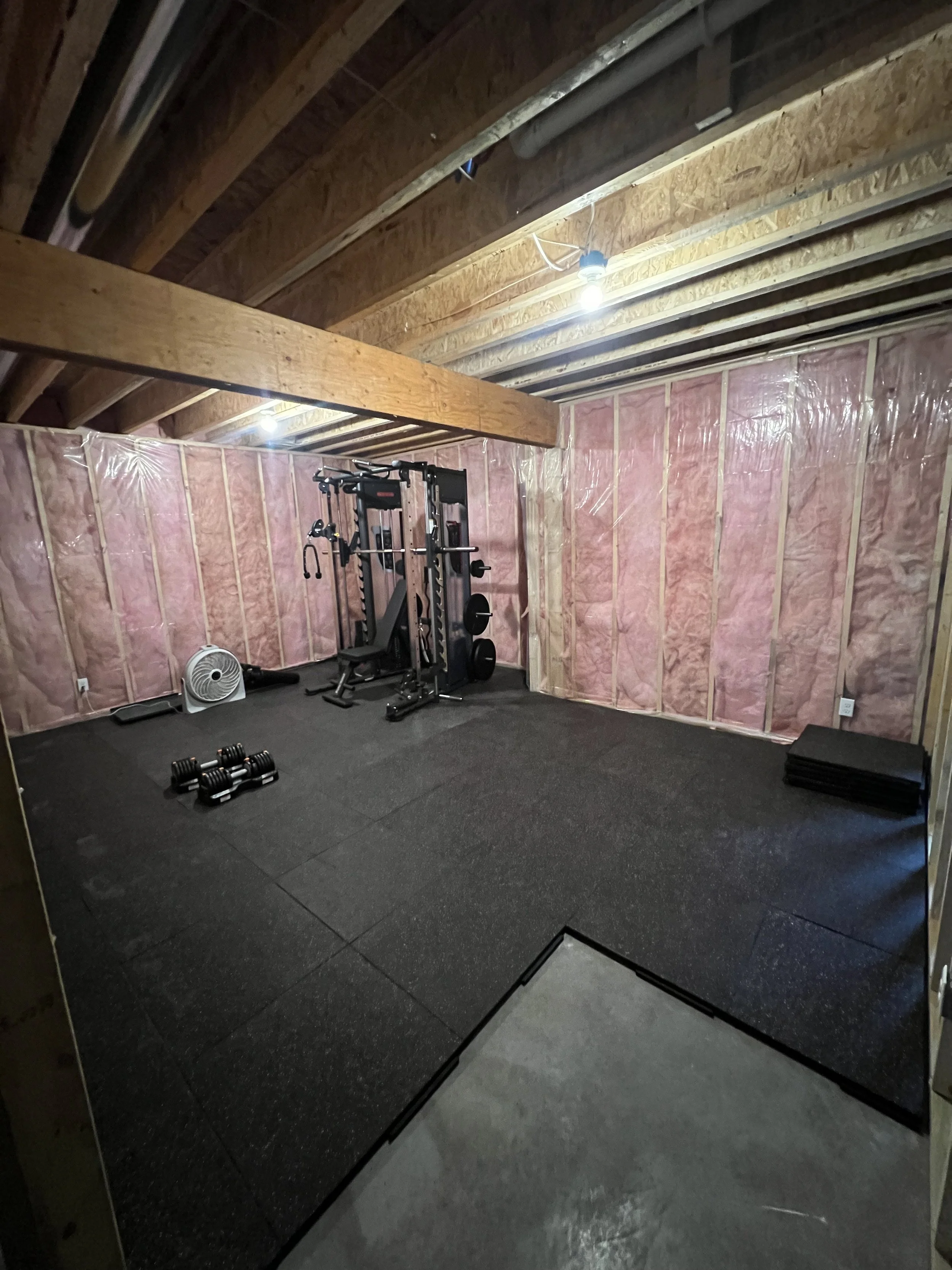  unfinished basement turned home gym with black rubber flooring, gym equipment, dumbbells, stacked weights, and pink insulation on the walls