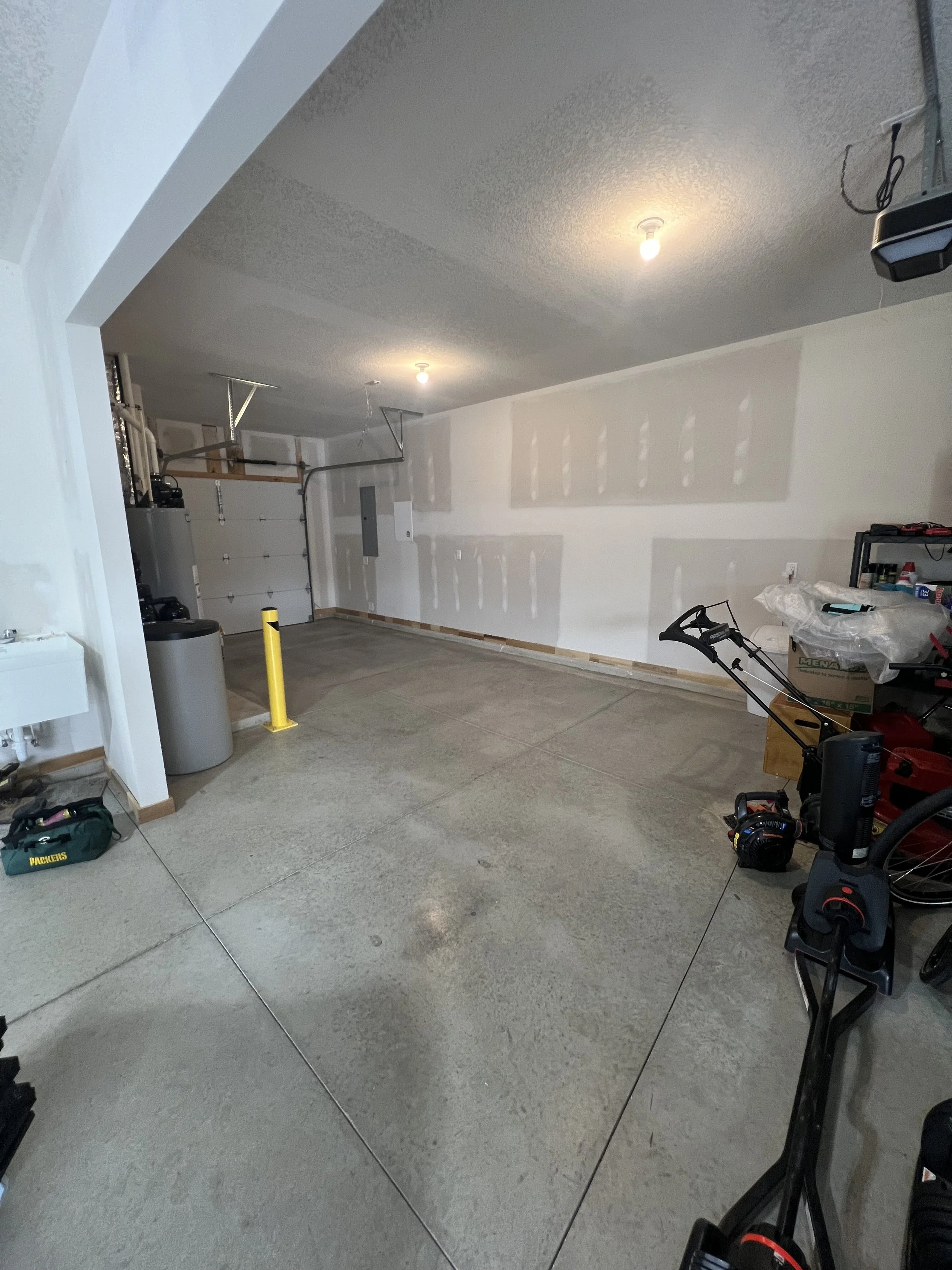 An unfinished garage with drywall on the walls, two light bulbs overhead, and various tools and equipment around the space.