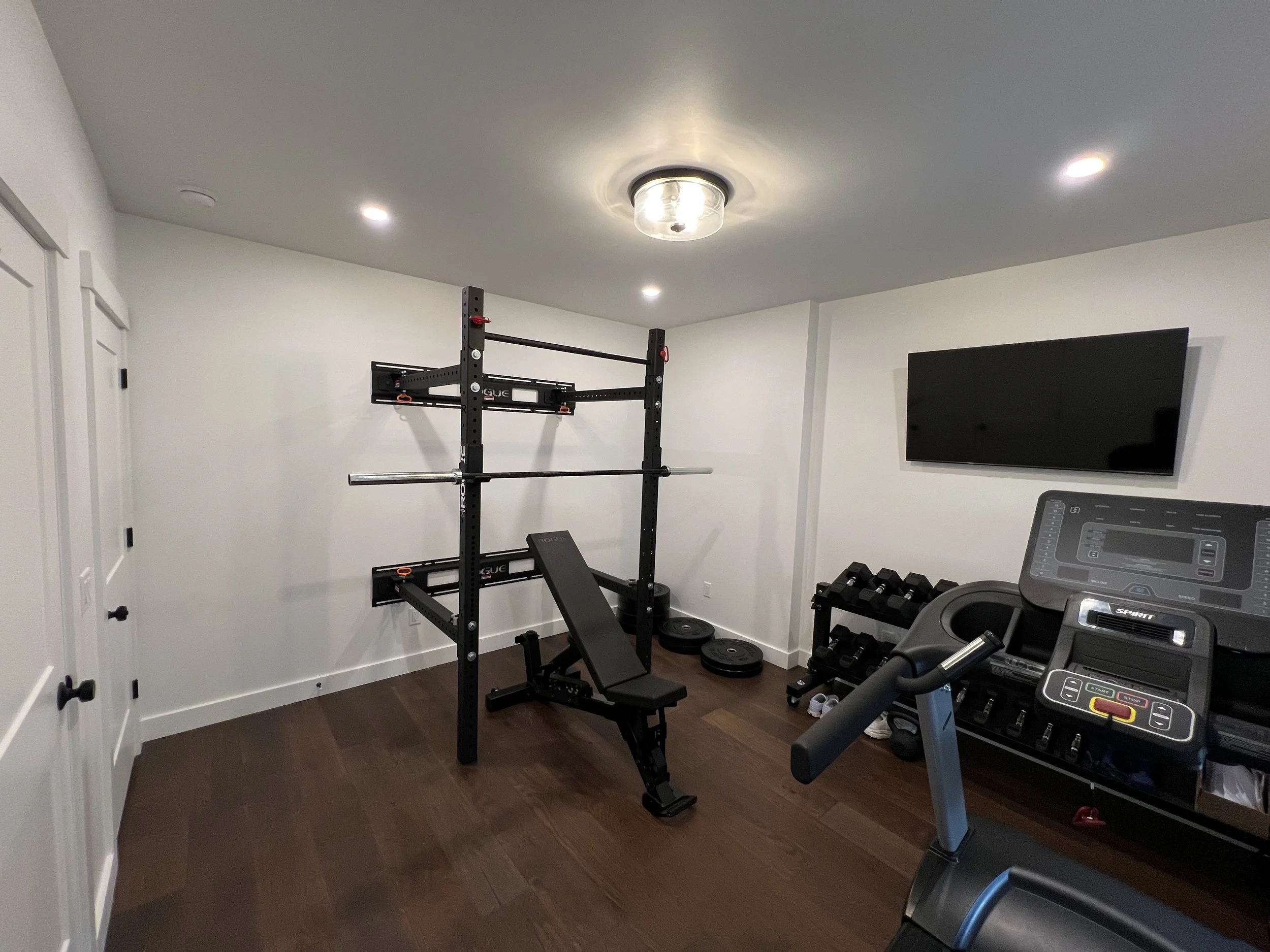 Home gym with a weightlifting squat rack with bars, a flat bench, dumbbells, weights, a treadmill, and a mounted flat-screen TV on a white wall.