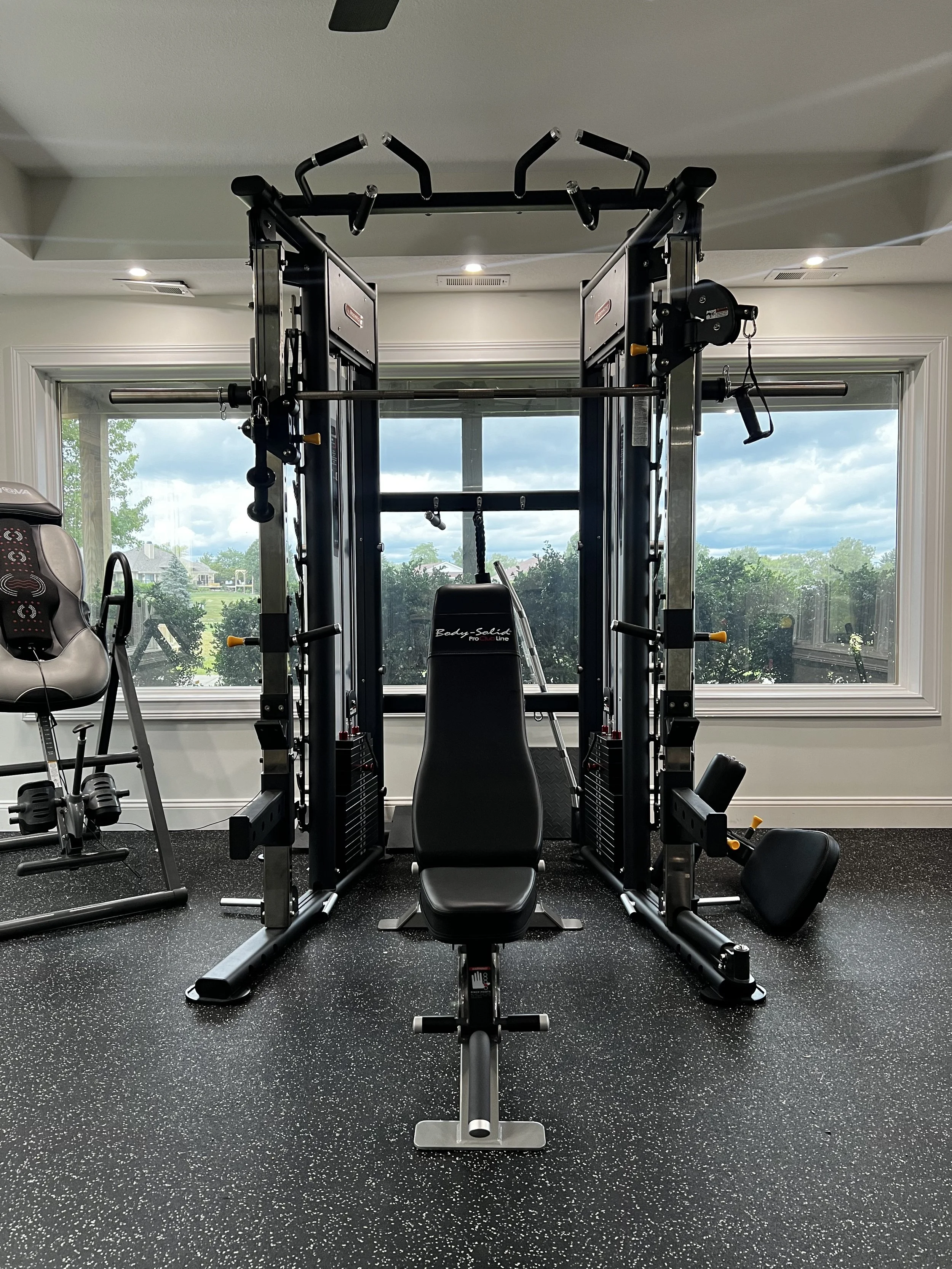 A home gym setup with a large black multi-station workout machine, a beige exercise chair, and a black workout bench on a speckled black rubber floor. Large windows in the background reveal a green, suburban landscape with trees and houses under a pa