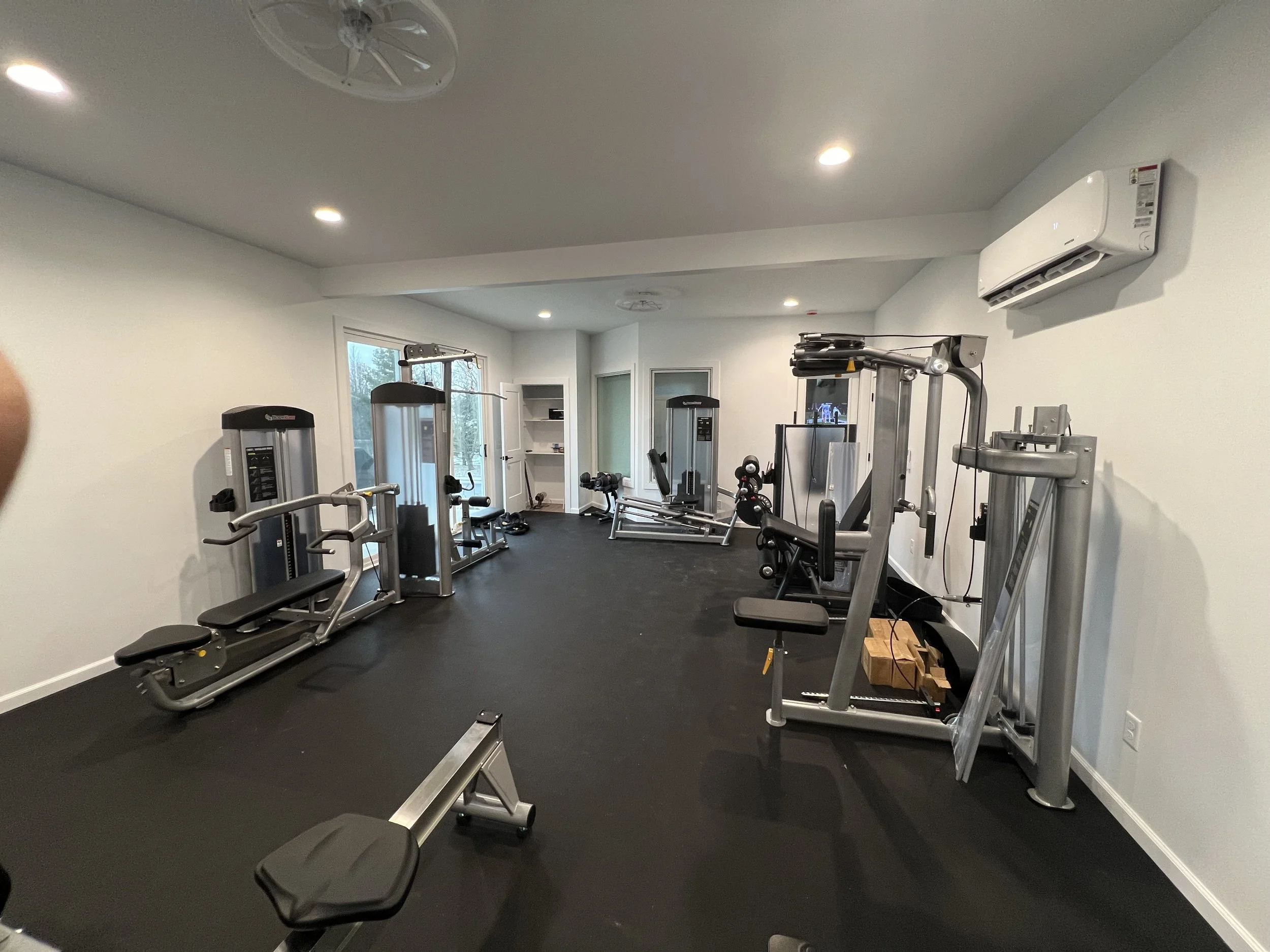 A home gym with various exercise equipment, including weight machines, a bench, and free weights, with white walls and a black rubber floor.