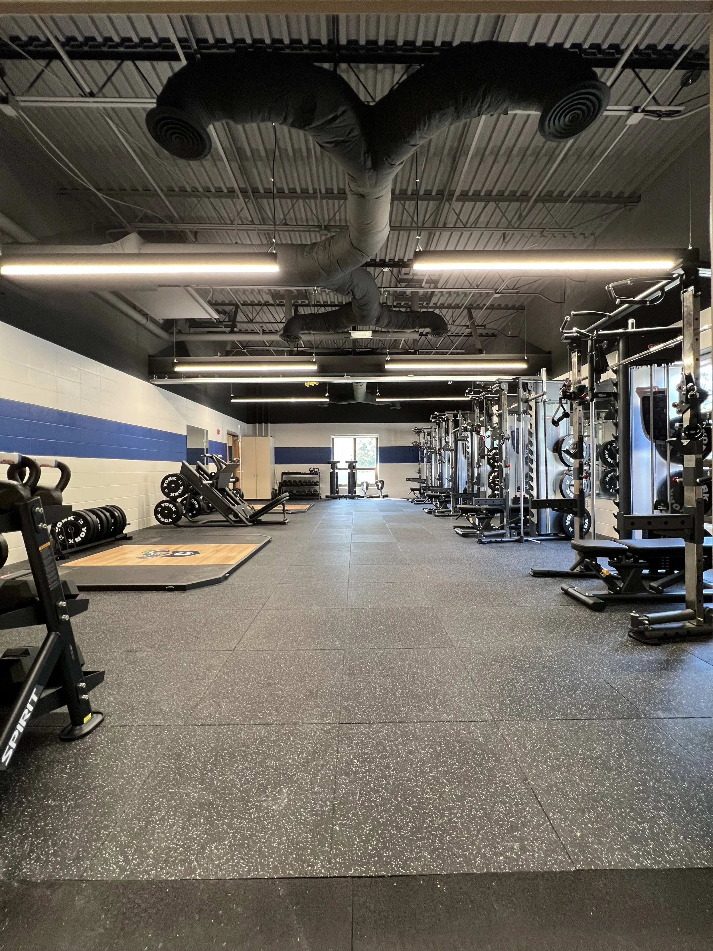 Empty gym with weightlifting equipment, benches, and squat racks, illuminated by fluorescent lights, with a window at the back.