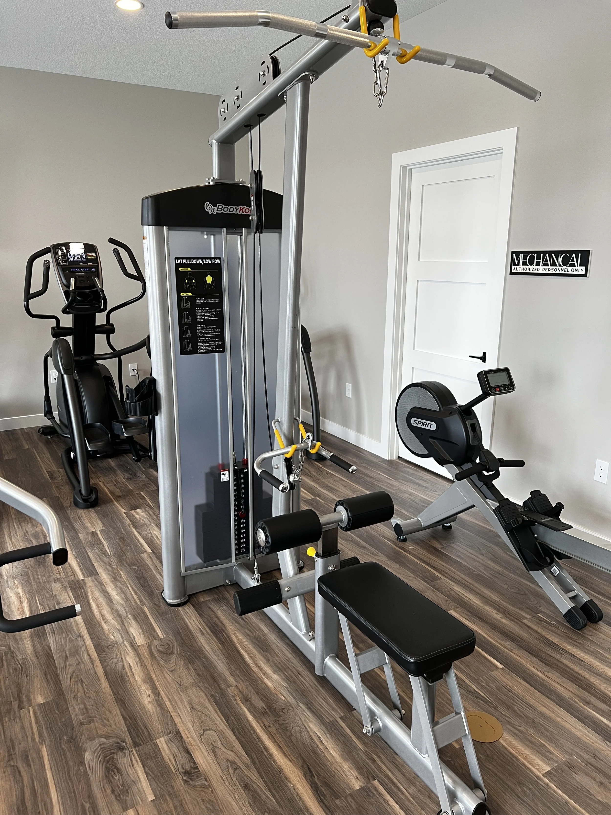 A home gym with various exercise equipment including a weight machine, an elliptical, a recumbent bike, and a workout bench, with a sign on the wall that says "Mechanical - Authorized Personnel Only."