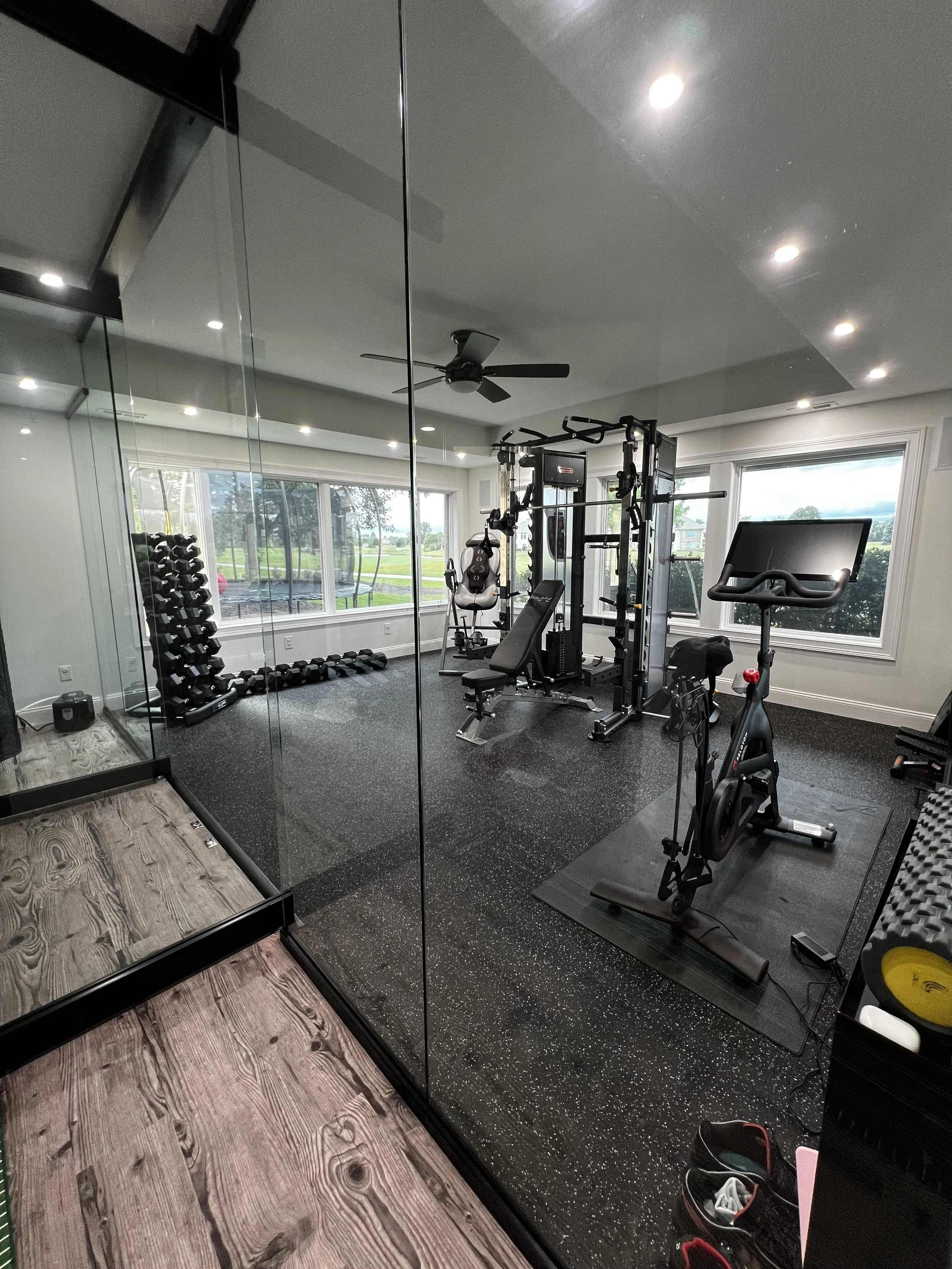 Modern home gym with various exercise equipment, including a treadmill, rowing machine, weightlifting station, and dumbbells, separated by glass walls, with windows showing greenery outside.
