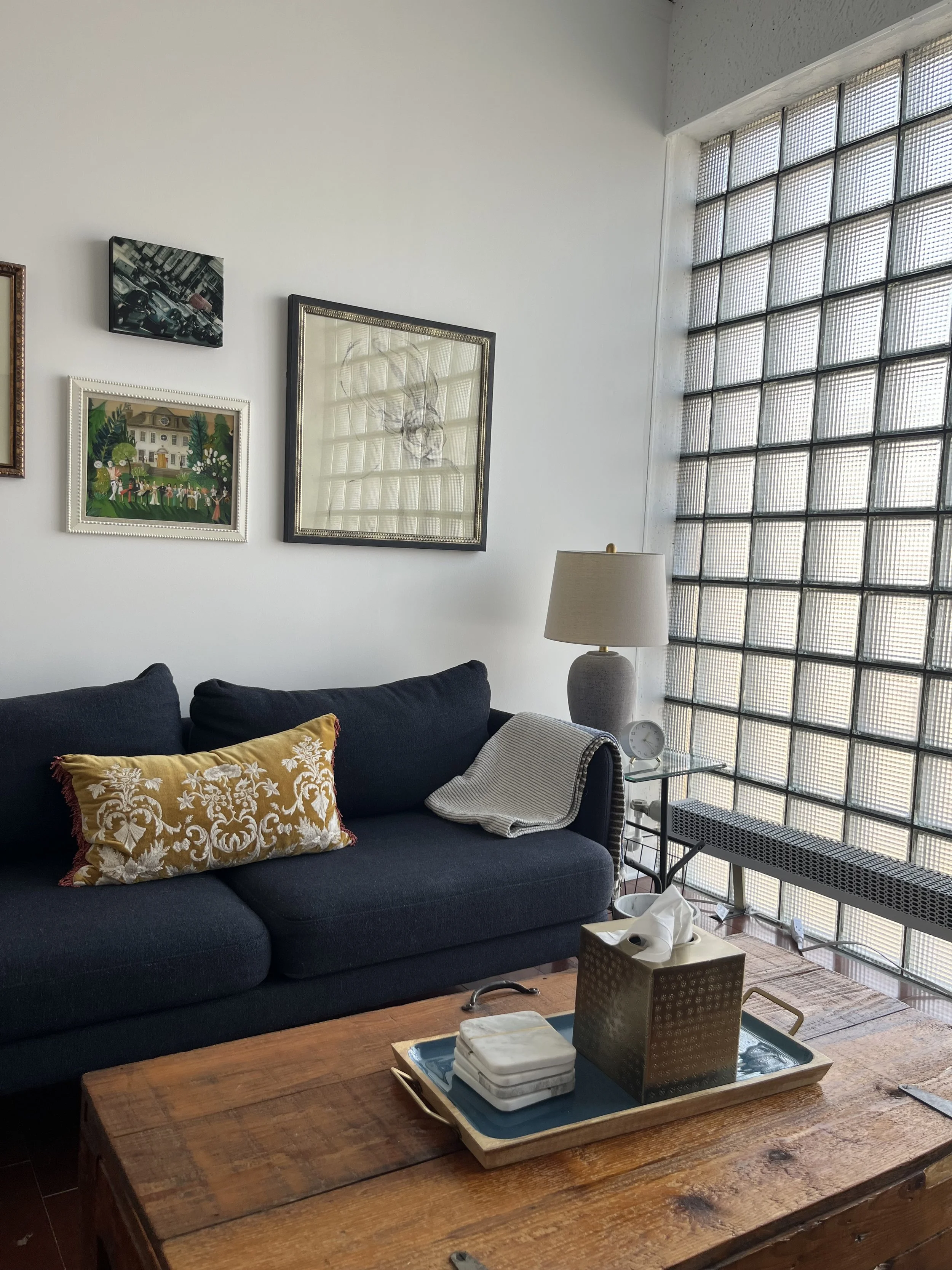 A therapist room with a dark blue sofa with a decorative yellow pillow and a striped throw blanket, a wooden coffee table with a tissue box and coaster, a glass side table with a lamp, and an abstract art wall with framed art pieces.