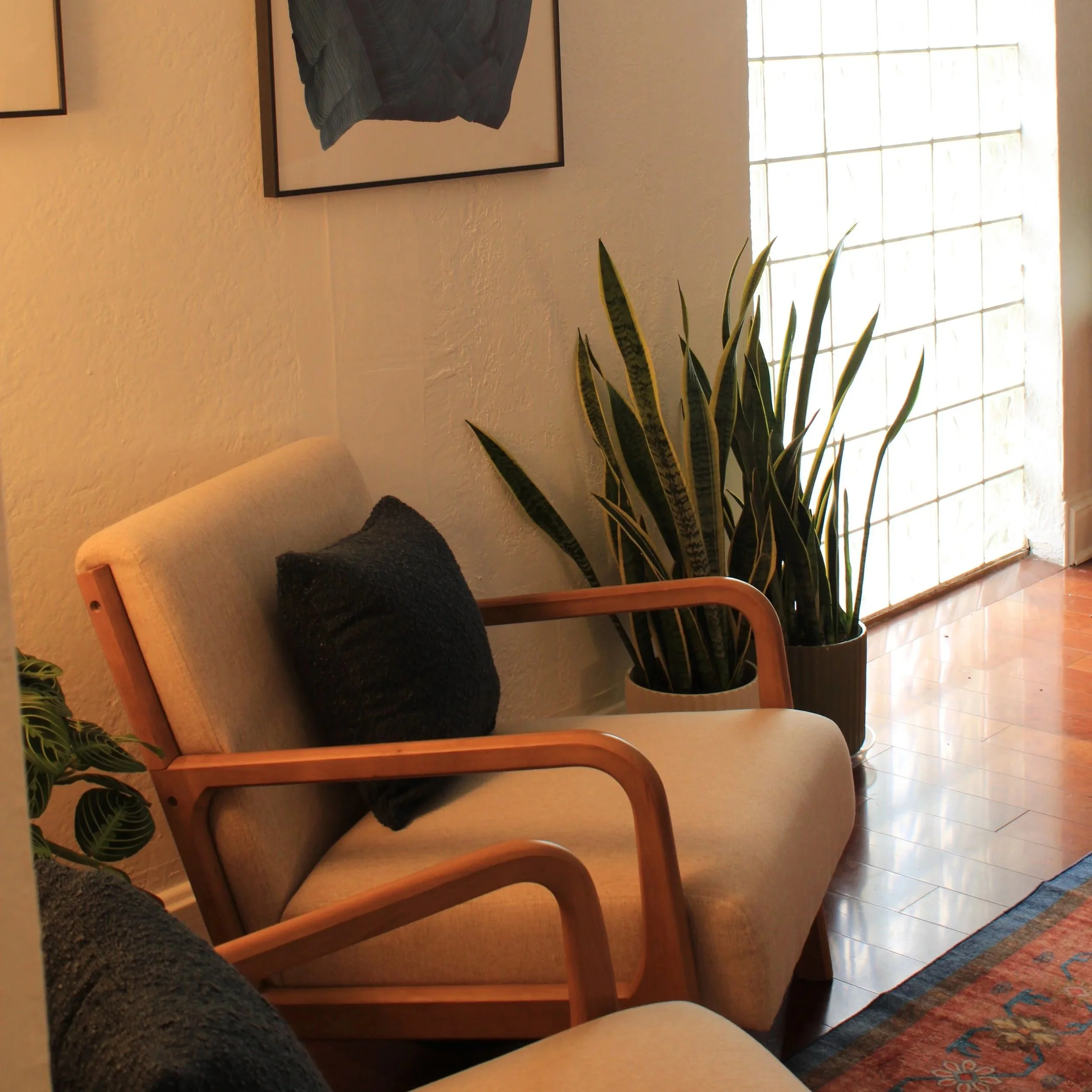 Corner of a cozy room with two beige chairs with wooden arms, a dark cushion on one chair, tall houseplants in black pots, a framed abstract artwork on the wall, a glass block window letting in natural light, and part of a colorful rug on a wooden fl