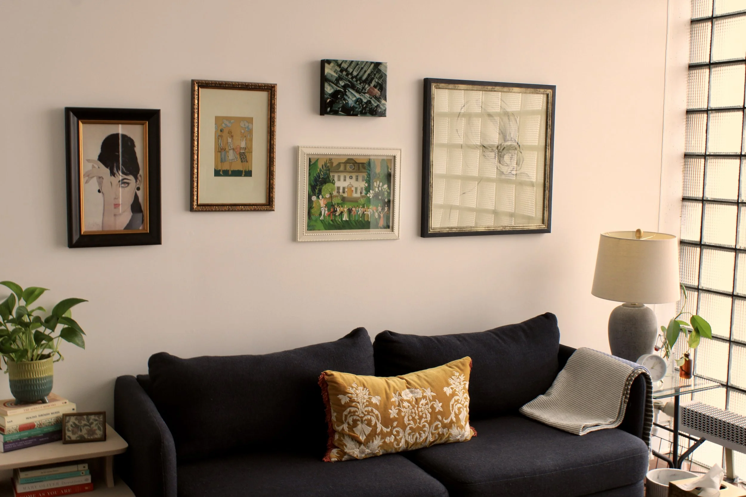 A therapy office with dark gray sofa, yellow decorative pillow, beige blanket, side table with books, potted plant, and paintings on white wall, with a large window on the right side.