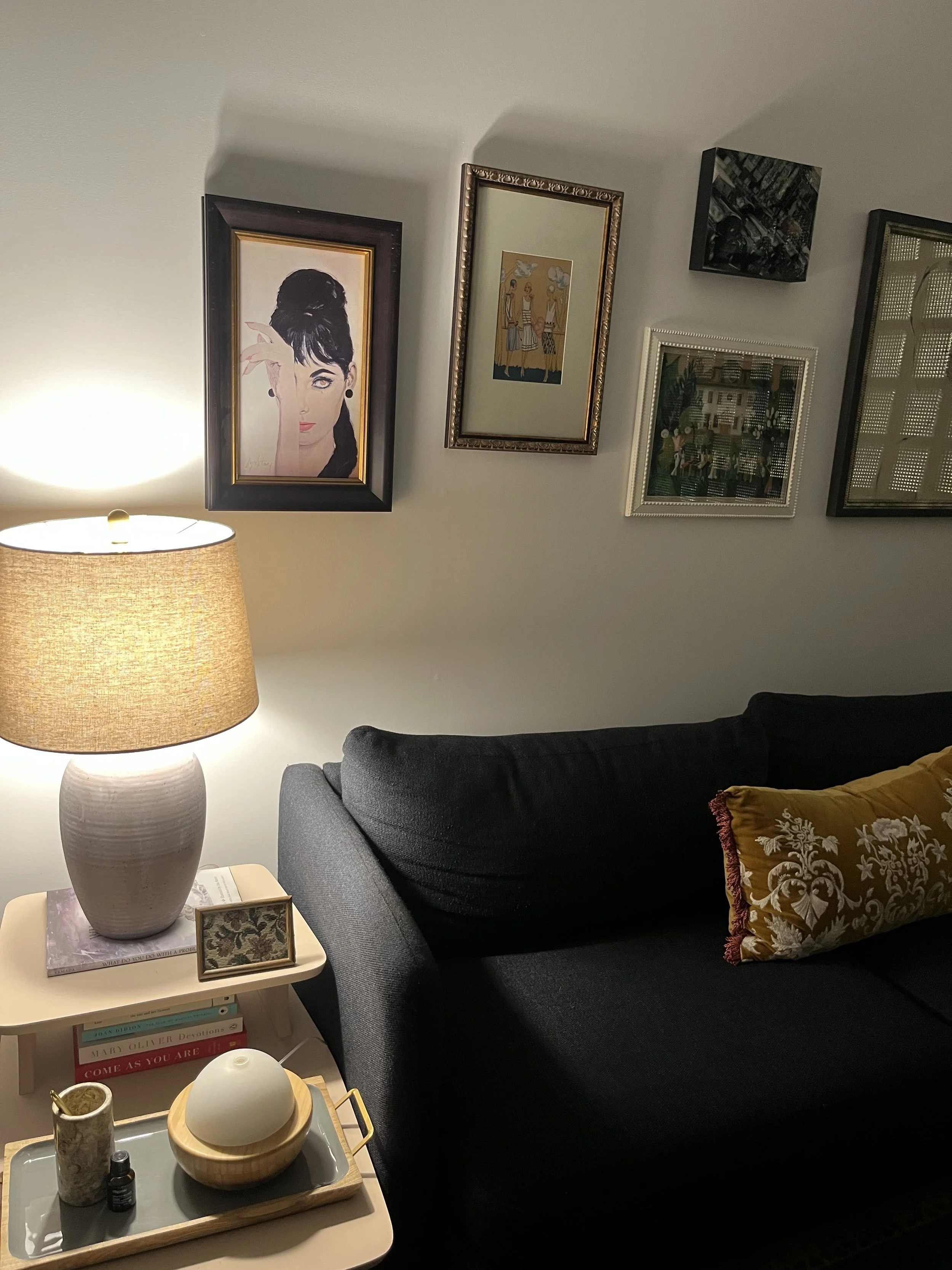 A therapy office with a dark gray sofa, beige side table, lamp, and multiple framed pictures on the wall. The side table has books, a small tray with a white object, a small bottle, and a decorative cup.
