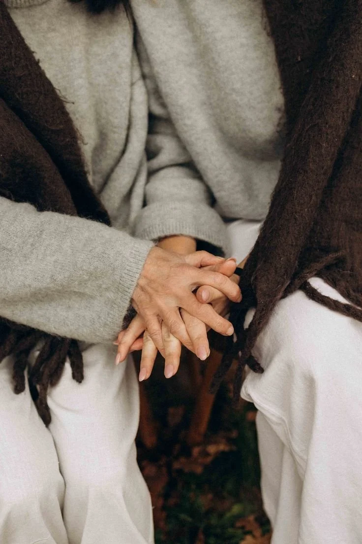 Two people holding hands, sitting outdoors on a fall day, with one wearing a gray sweater and the other a brown jacket.