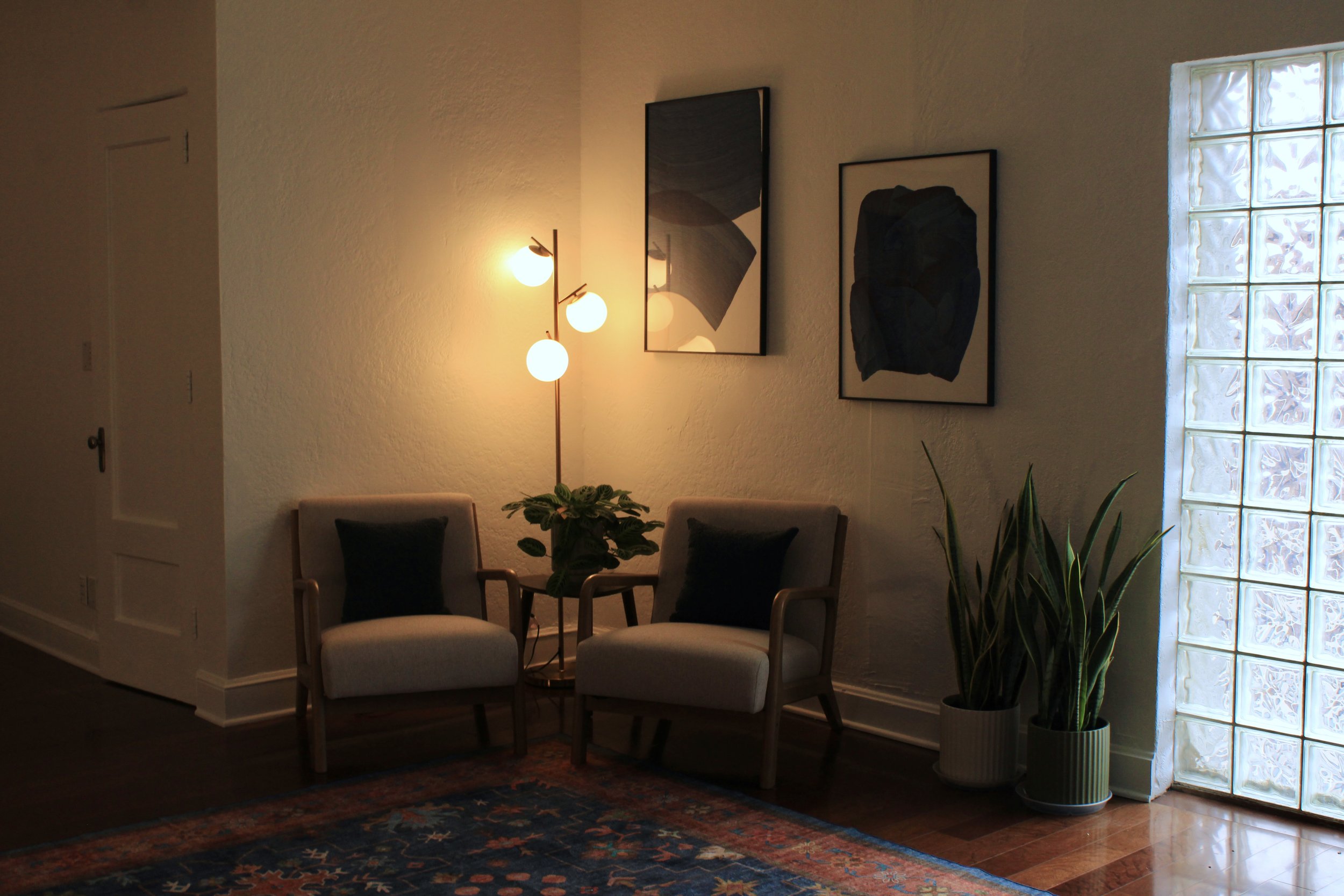A cozy interior living space with two beige armchairs with dark pillows, a glass block window, a potted snake plant, a small table with a leafy plant, a tall floor lamp with multiple round lights, and three framed abstract artworks on the wall.