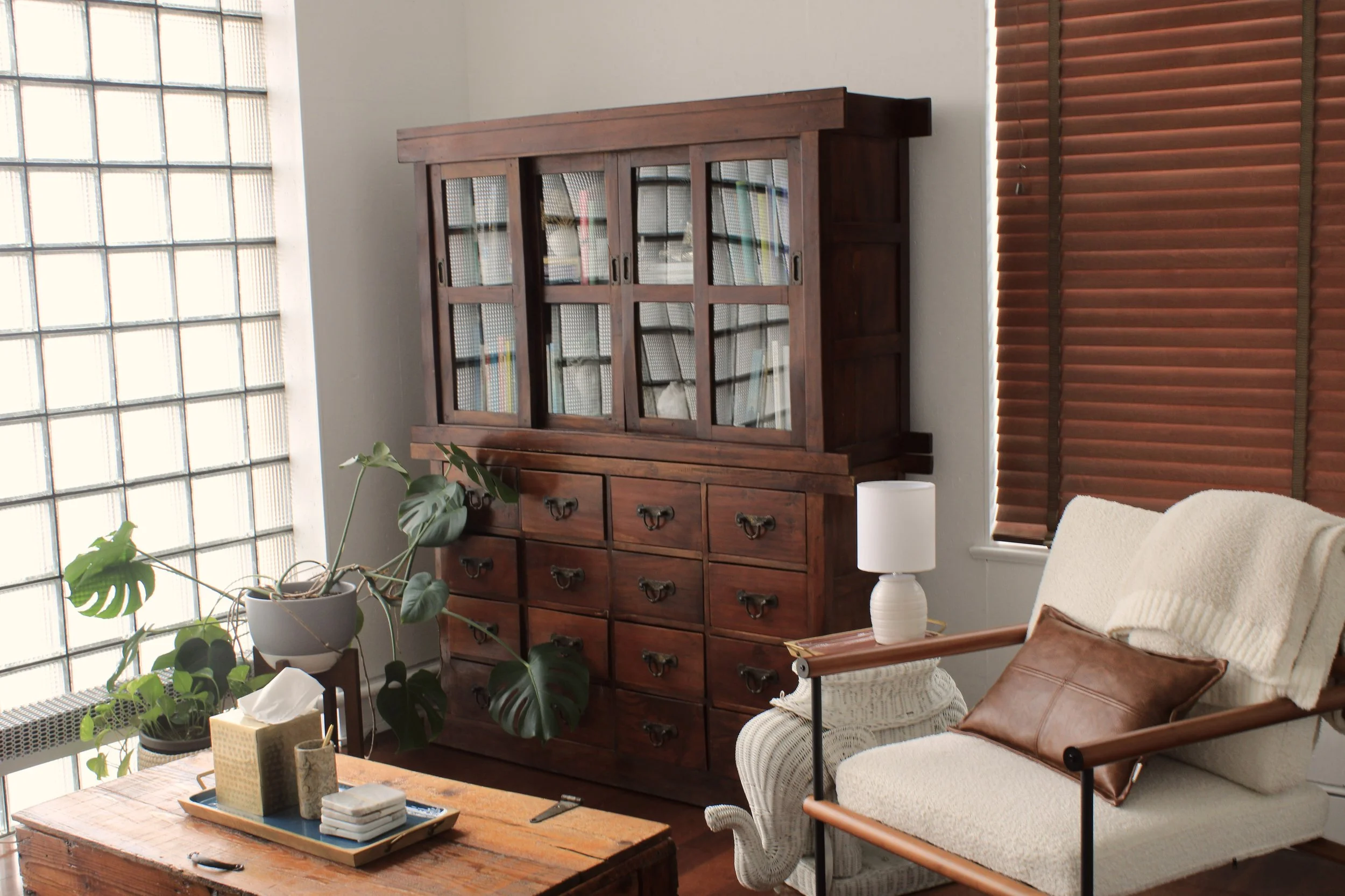 Interior of a therapist's office space with a wooden cabinet, a plant with large leaves, a table with a tray of items, a white lamp on an armchair, and brown blinds covering the windows.
