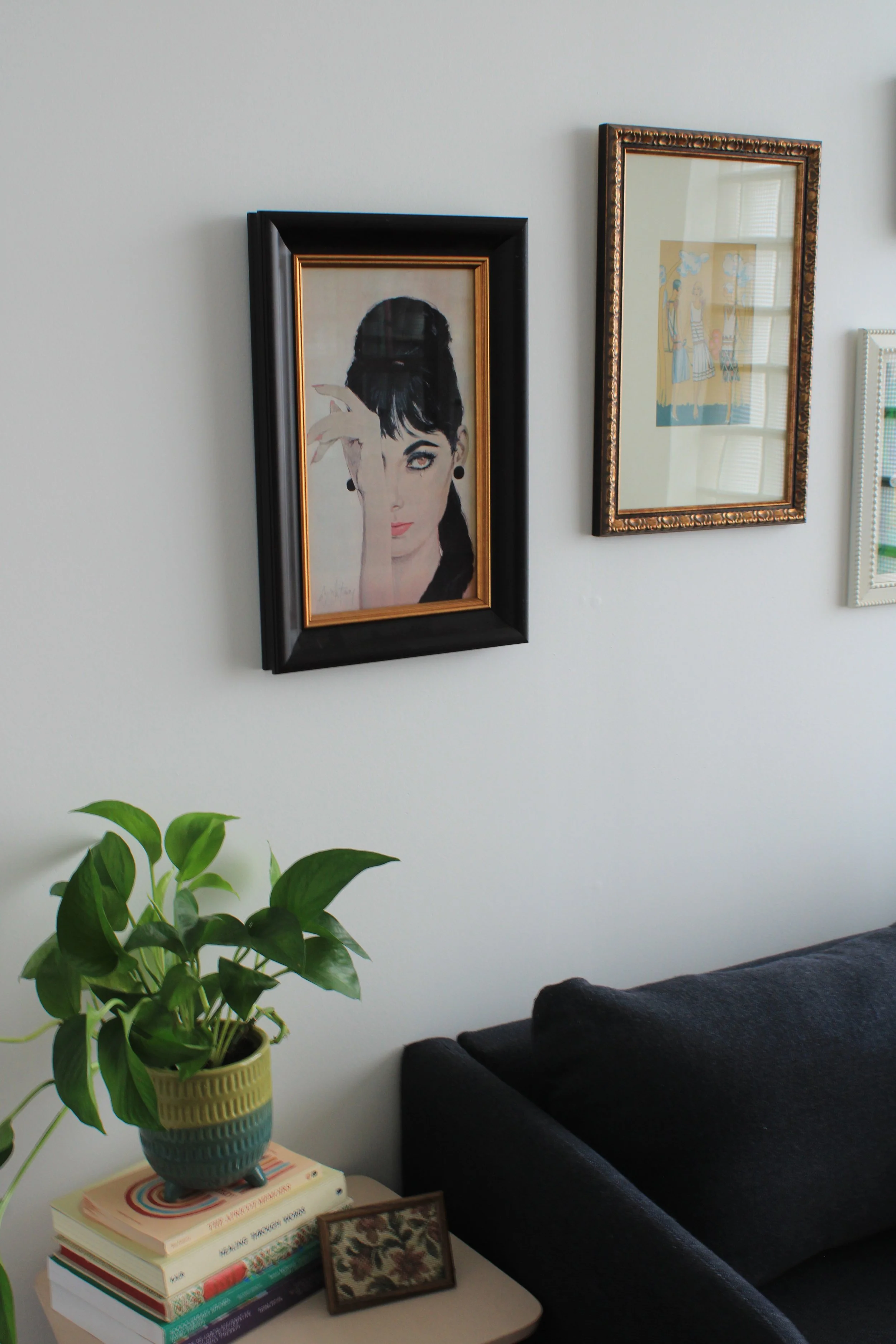 A white wall with two framed artwork pieces; one features a woman with dark hair, partly covering her face with her hand, and the other depicts a scene with people. Below the art, there's a white table with a stack of books, a framed picture, and a potted green plant. A black sofa is partially visible in the foreground.