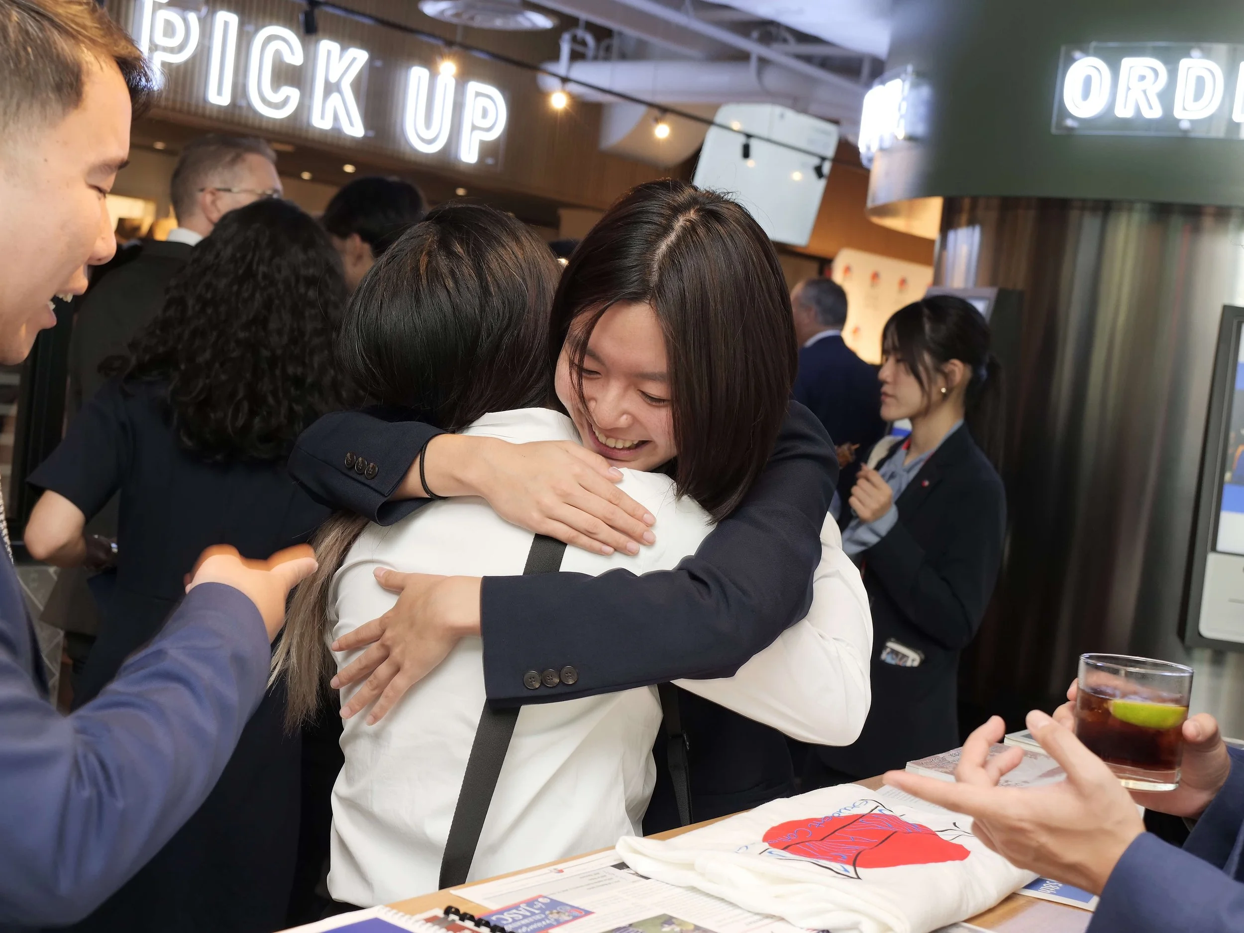 Two ISC delegates share a warm hug during a conference closing event, surrounded by fellow participants in conversation.
