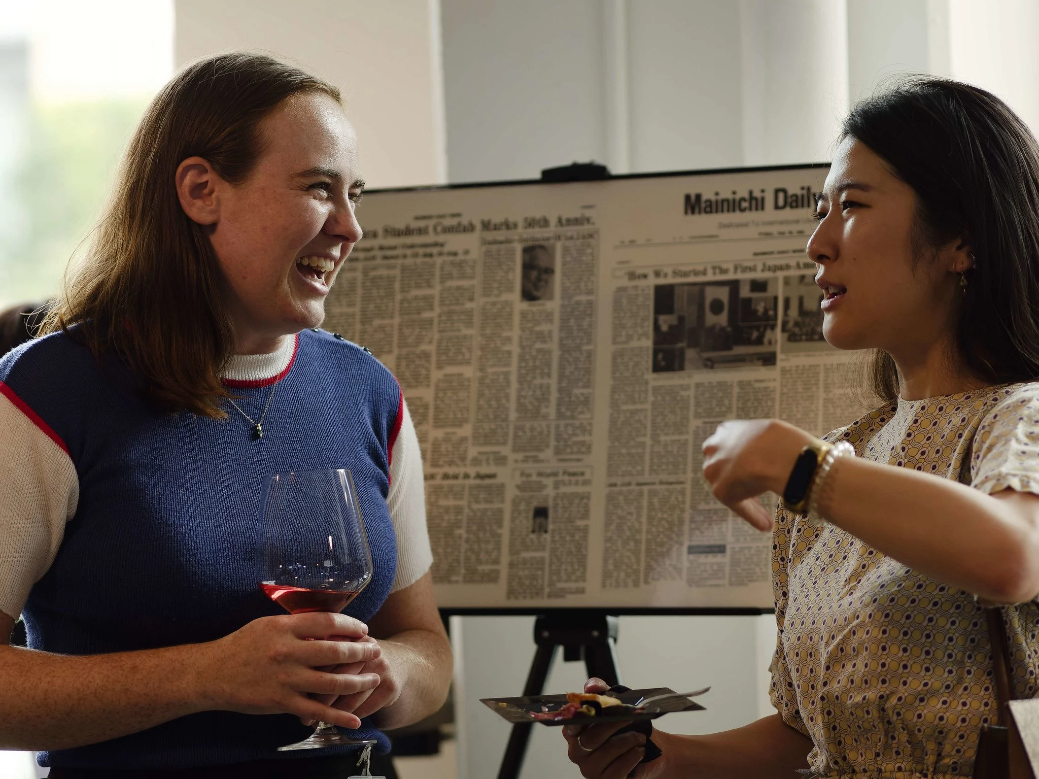 Two ISC alumni smile and talk during a networking reception, with a historic Japan–America Student Conference newspaper displayed in the background.