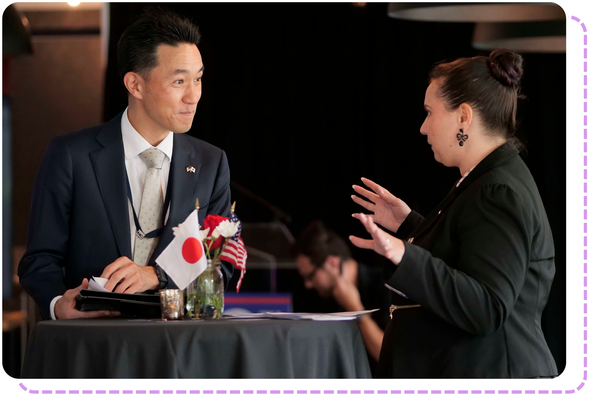 Two people in professional attire engage in a lively conversation at a table decorated with small Japanese and American flags during an International Student Conferences event.