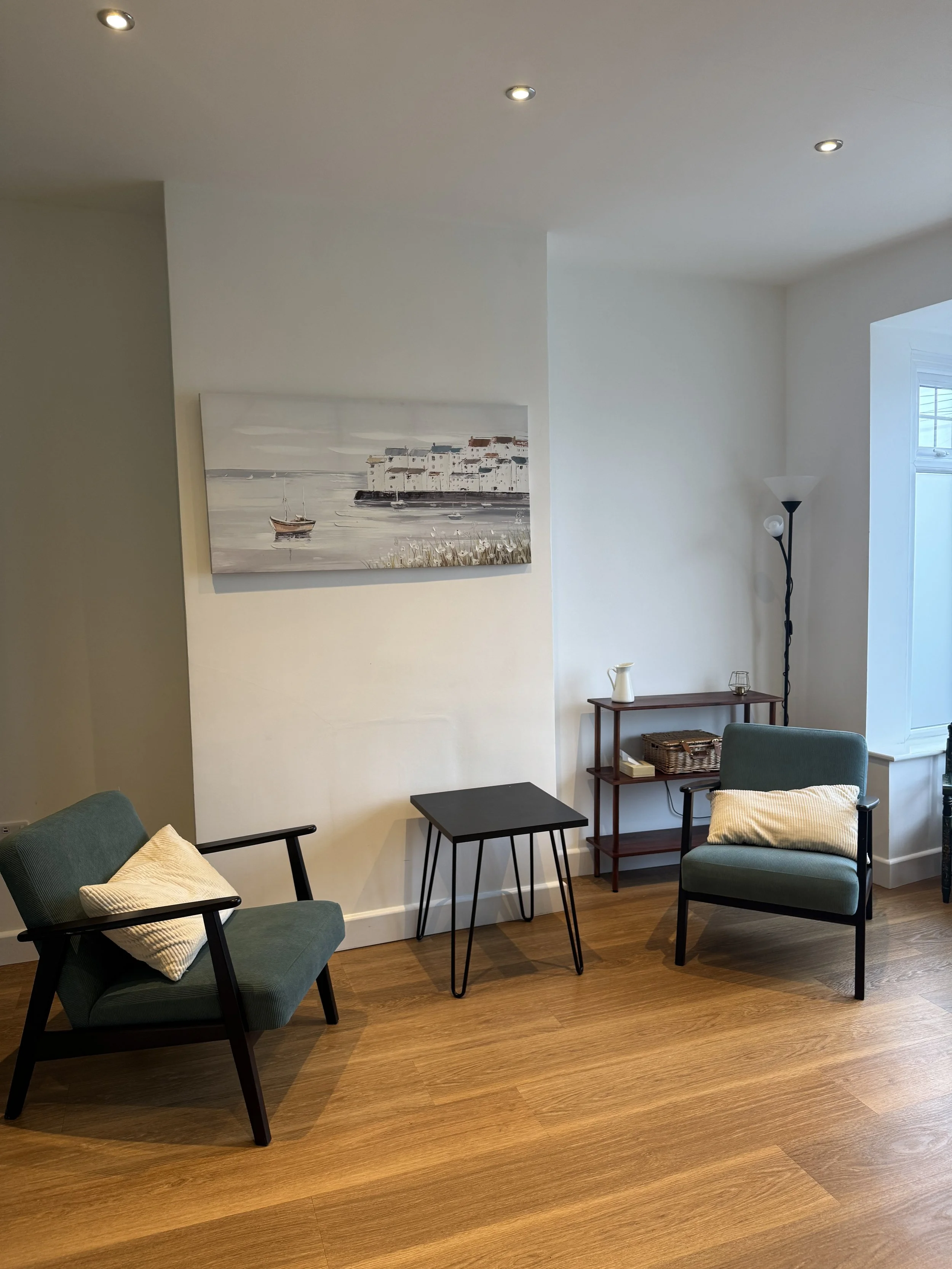 A cozy living room with two green armchairs with light-colored pillows, a small black side table, a wooden shelving unit with decorative items, a tall floor lamp, a window, and a landscape painting of a coastal scene, all on hardwood flooring.