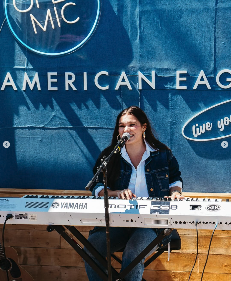 A woman smiling and singing into a microphone while playing a Yamaha keyboard during a live performance at an American Eagle event.