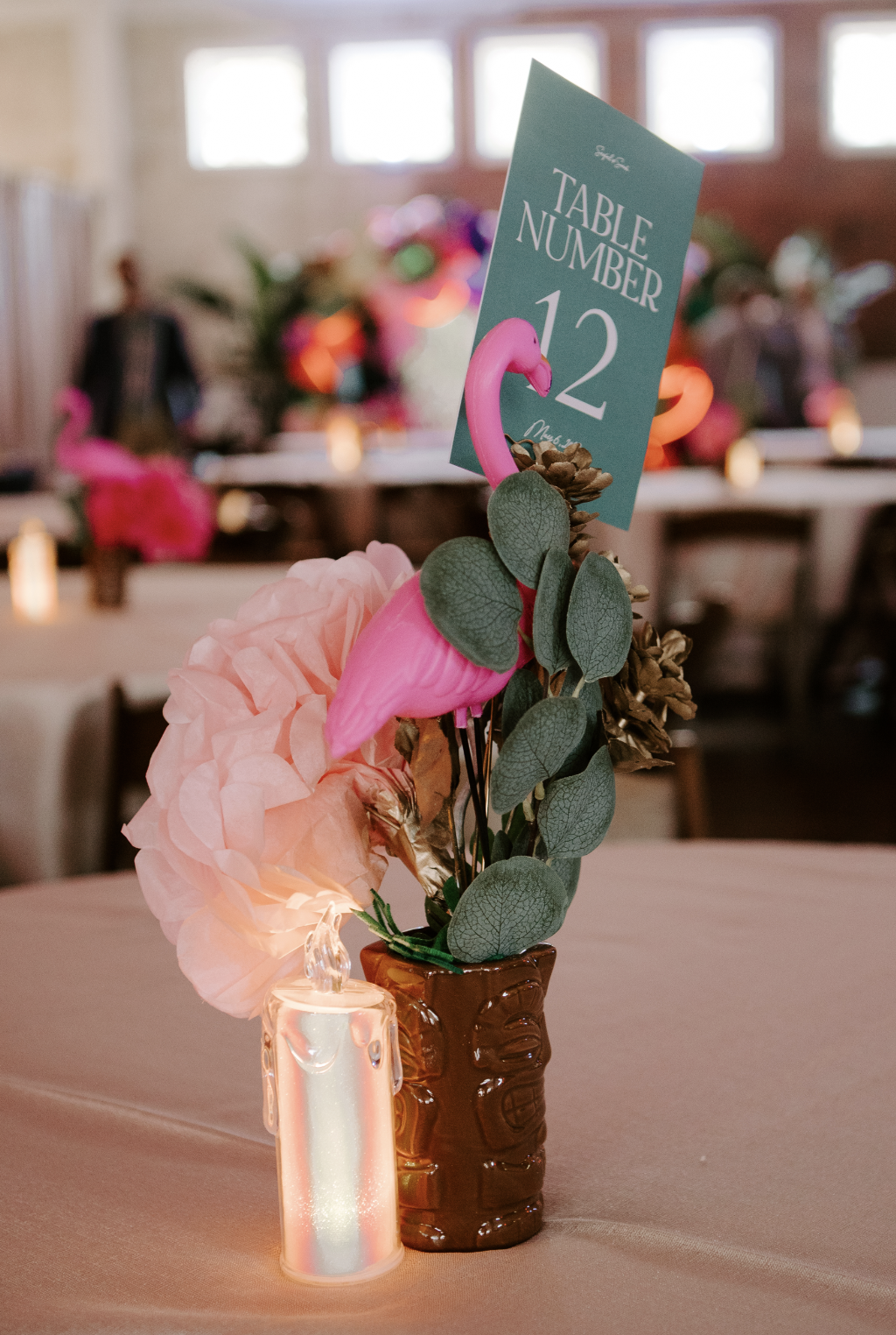 Centerpiece with pink flamingo, pink tissue paper flowers, eucalyptus leaves, and a table number sign reading 'Table Number 12' in a decorated event venue.