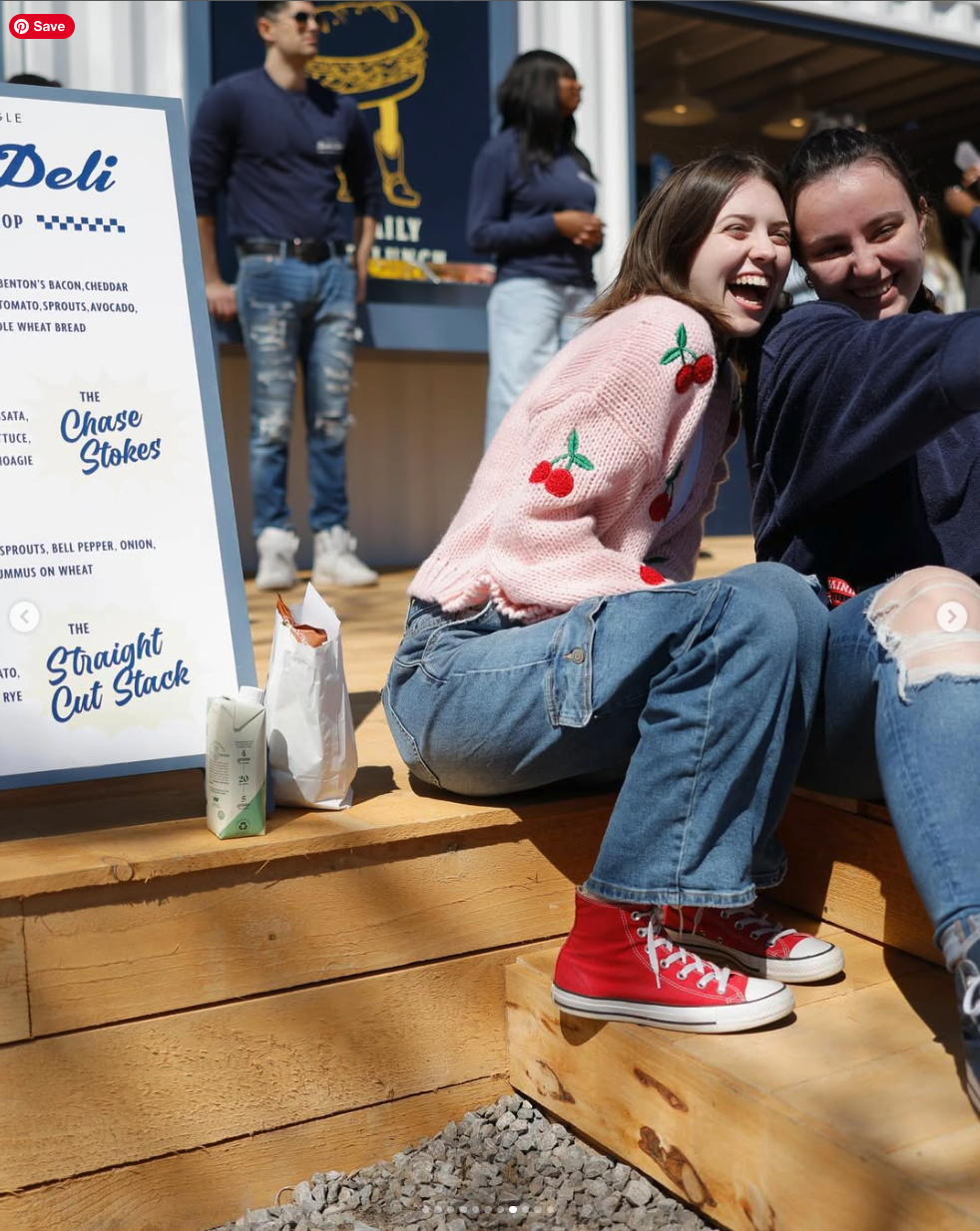 Two young women are sitting and smiling at the camera in an outdoor setting, with a menu board and people in the background. One woman is wearing a pink sweater with cherry designs, ripped jeans, and red sneakers. The other is wearing a dark shirt and jeans.