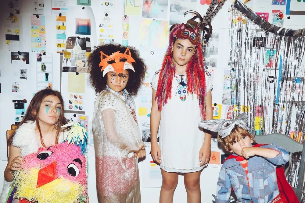 Four children dressed in costumes and face paint, standing in front of a colorful mural or collage with artwork and posters, with decorations including tinsel on the right side.