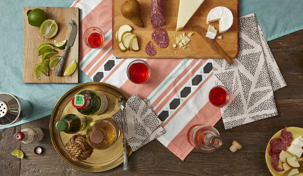Flat lay of cheese, sliced meats, limes, and beverages on a wooden table with patterned napkins and a colorful tablecloth.
