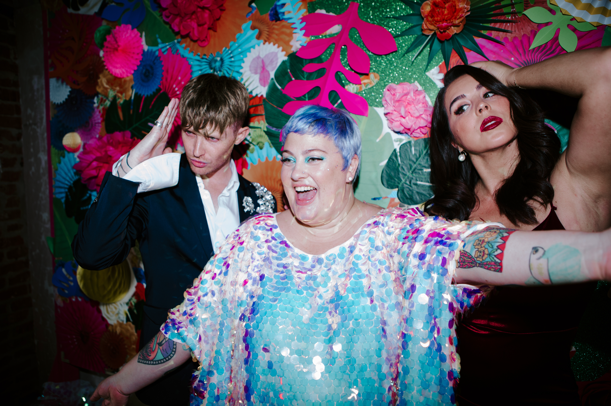 Three people at a colorful, festive party pose playfully against a vibrant background of oversized paper flowers and tropical leaves. The person in the center has short light purple hair, is smiling broadly, and wearing a shiny, iridescent top. The person on the left has short brown hair, is wearing a dark jacket with embellishments, and is making a gesture near their head. The person on the right has long dark hair, wears bold makeup, and is wearing a dark dress with a tattoo on their arm.