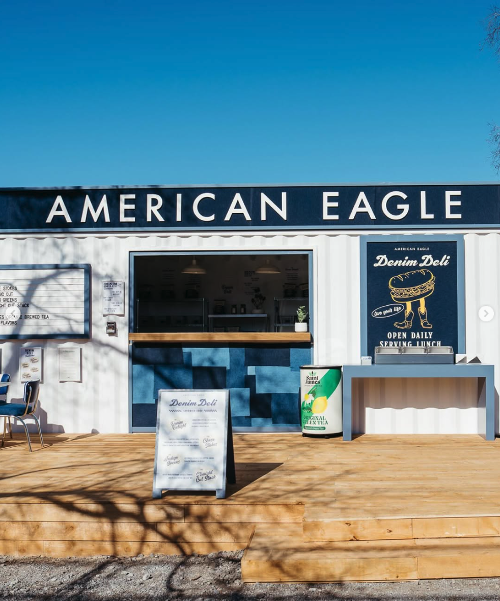 Front view of a food stand named American Eagle with a sign for Denim Deli, featuring a menu board, sign for original green tea, and outdoor seating area on a wooden deck.