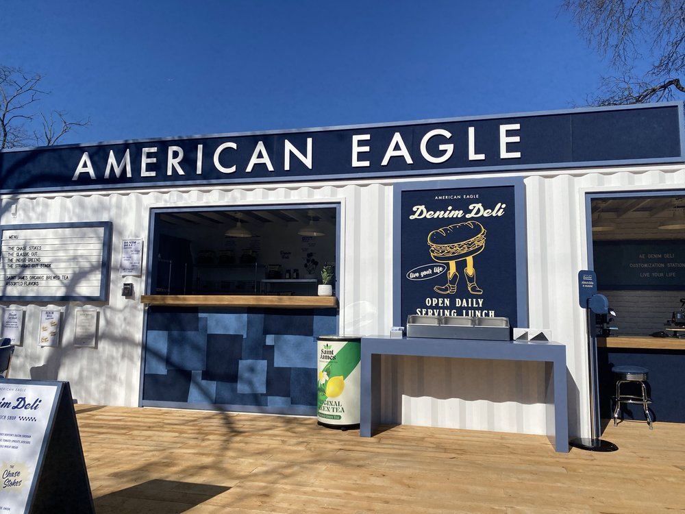Food truck with sign 'American Eagle' and a logo for 'Denim Deli' featuring a hot dog with legs and boots, outside on a wooden deck under a clear blue sky.