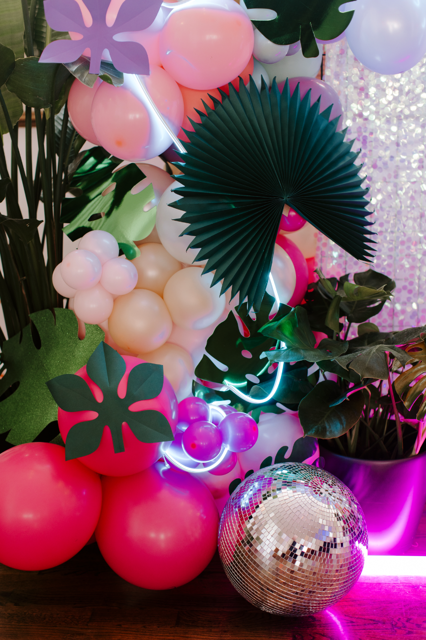 Decorative arrangement with pink, white, and green balloons, large green paper leaves, a black paper fan, a disco ball, and colorful neon lights, set against a pink sparkly background and potted green plants.