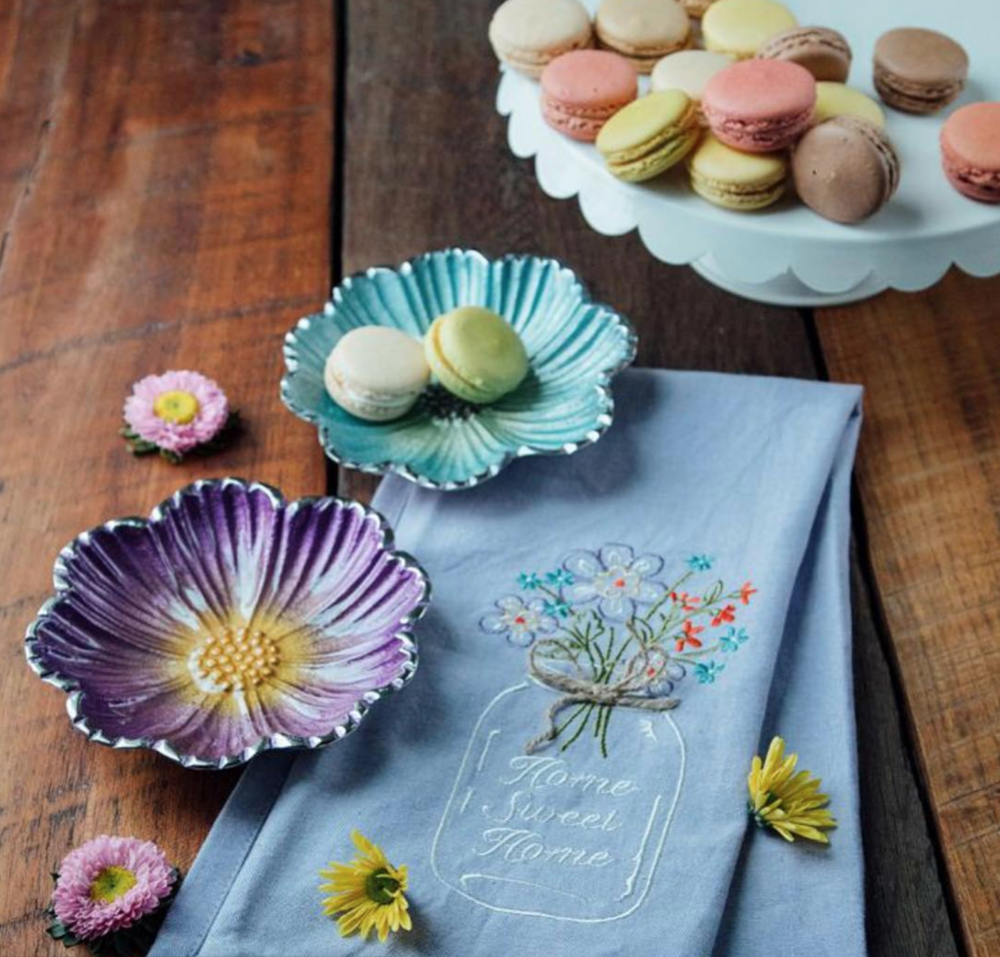 A table set with a blue embroidered cloth, decorative floral bowls, and a white cake stand with assorted macarons in pastel colors. Small flowers are scattered around, featuring pink, yellow, and purple flowers.