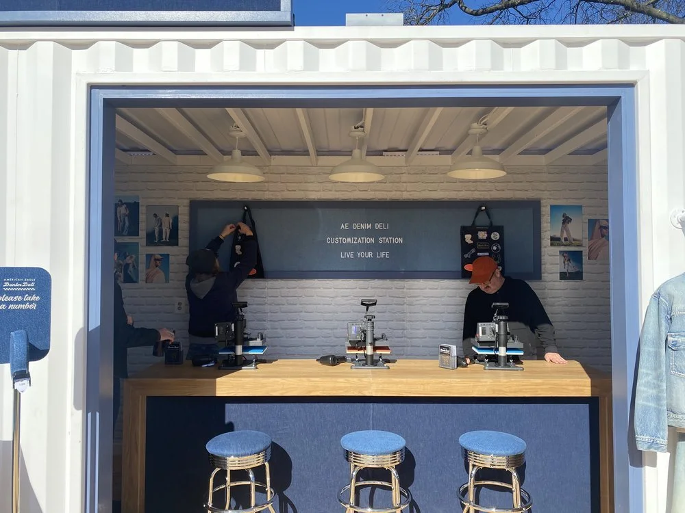 Inside a denim customization booth with two employees working on denim jackets, blue stools in front, and denim photos on the wall.