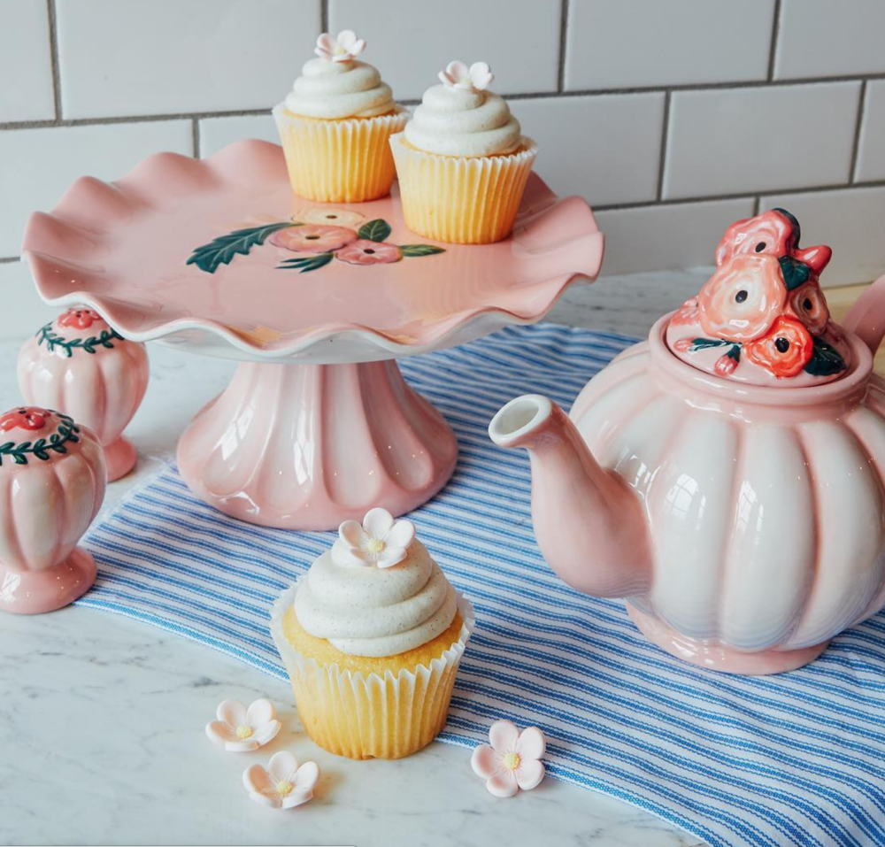Pink ceramic tea set with a teapot and cups, decorated with floral motifs, along with a pink cupcake with white frosting and a small flower decoration, displayed on a striped blue and white cloth on a marble surface.
