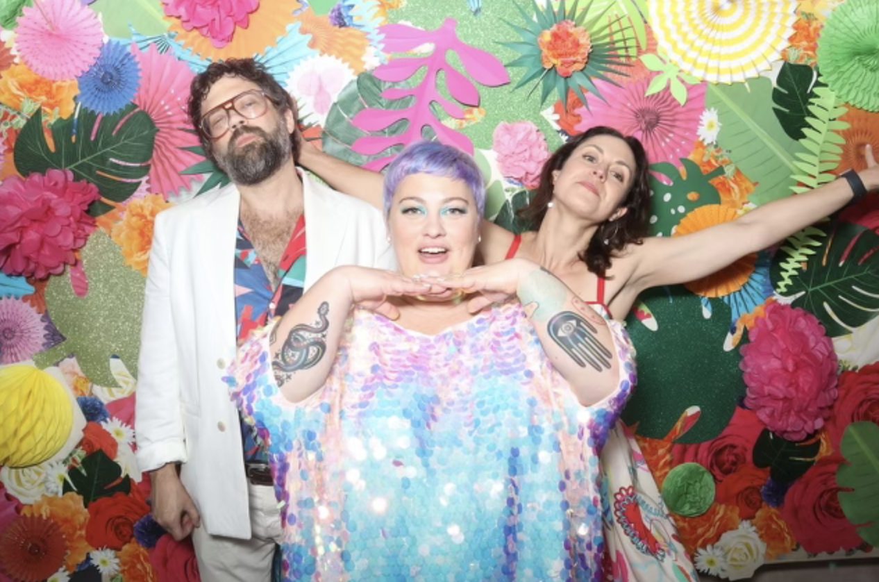 Three people pose in front of a colorful floral backdrop. The person in the center has short, pastel purple hair and is wearing a shiny, pastel, iridescent top, with tattoos on both arms. The person on the left has curly hair, glasses, a beard, and is wearing a white blazer over a vibrant shirt. The person on the right has shoulder-length dark hair, is wearing a red top, and has one arm outstretched. The background features large, colorful paper flowers and leaves.