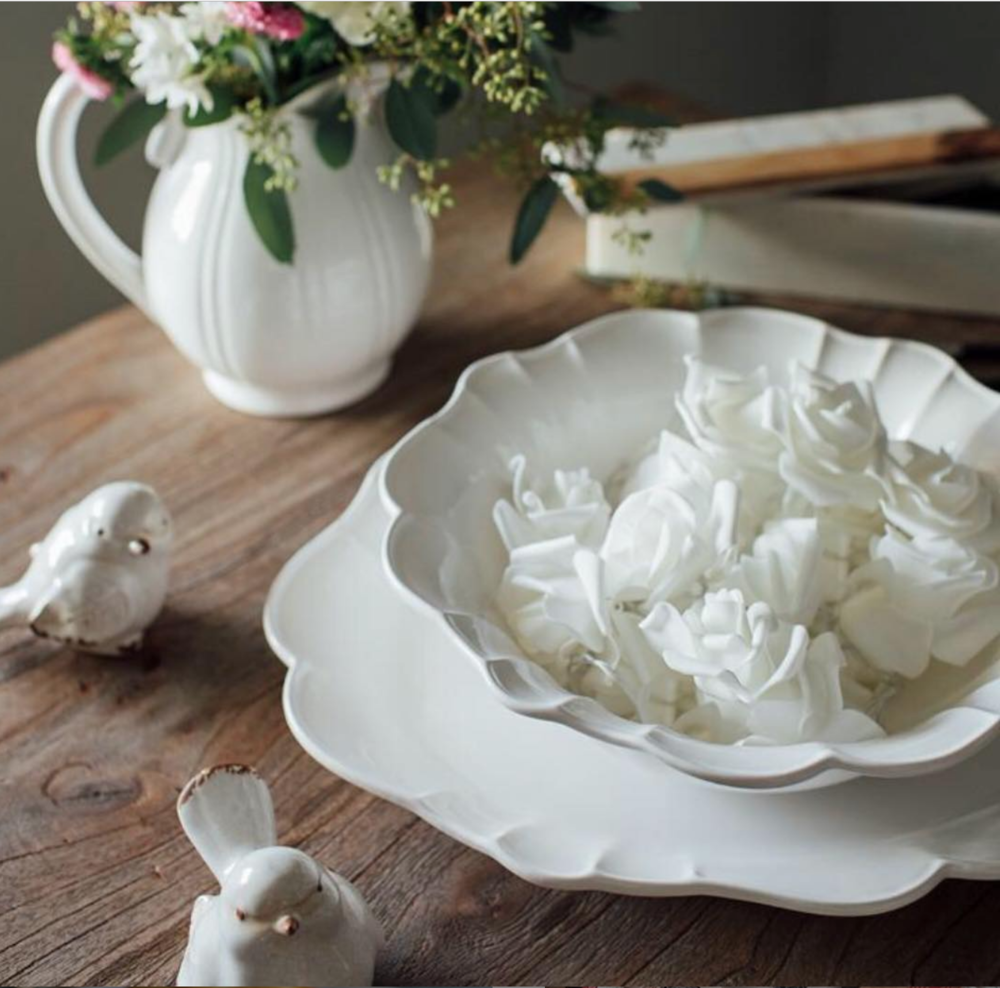 White ceramic bowl filled with white flower-shaped candies on a wooden table, accompanied by a white ceramic vase with pink and green floral arrangements and small ceramic bird figurines, with books stacked in the background.