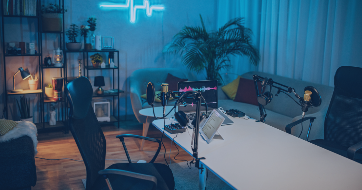 A podcast recording setup in a cozy, modern room with a white table, multiple microphones, headphones, a laptop, a tablet, and a sound mixer. The room has a sofa, a large plant, bookshelves, and decorative lighting, including a neon sign on the wall.