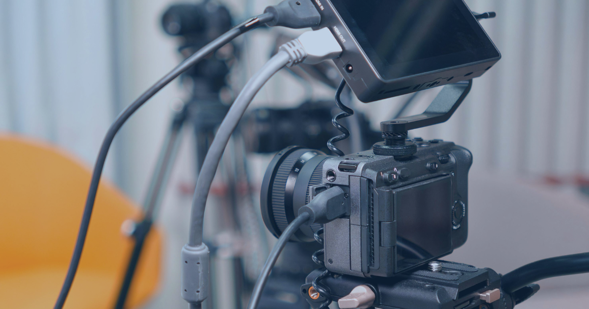Close-up of a professional camera setup with a monitor mounted above, various cables connected, and a tripod in the background.
