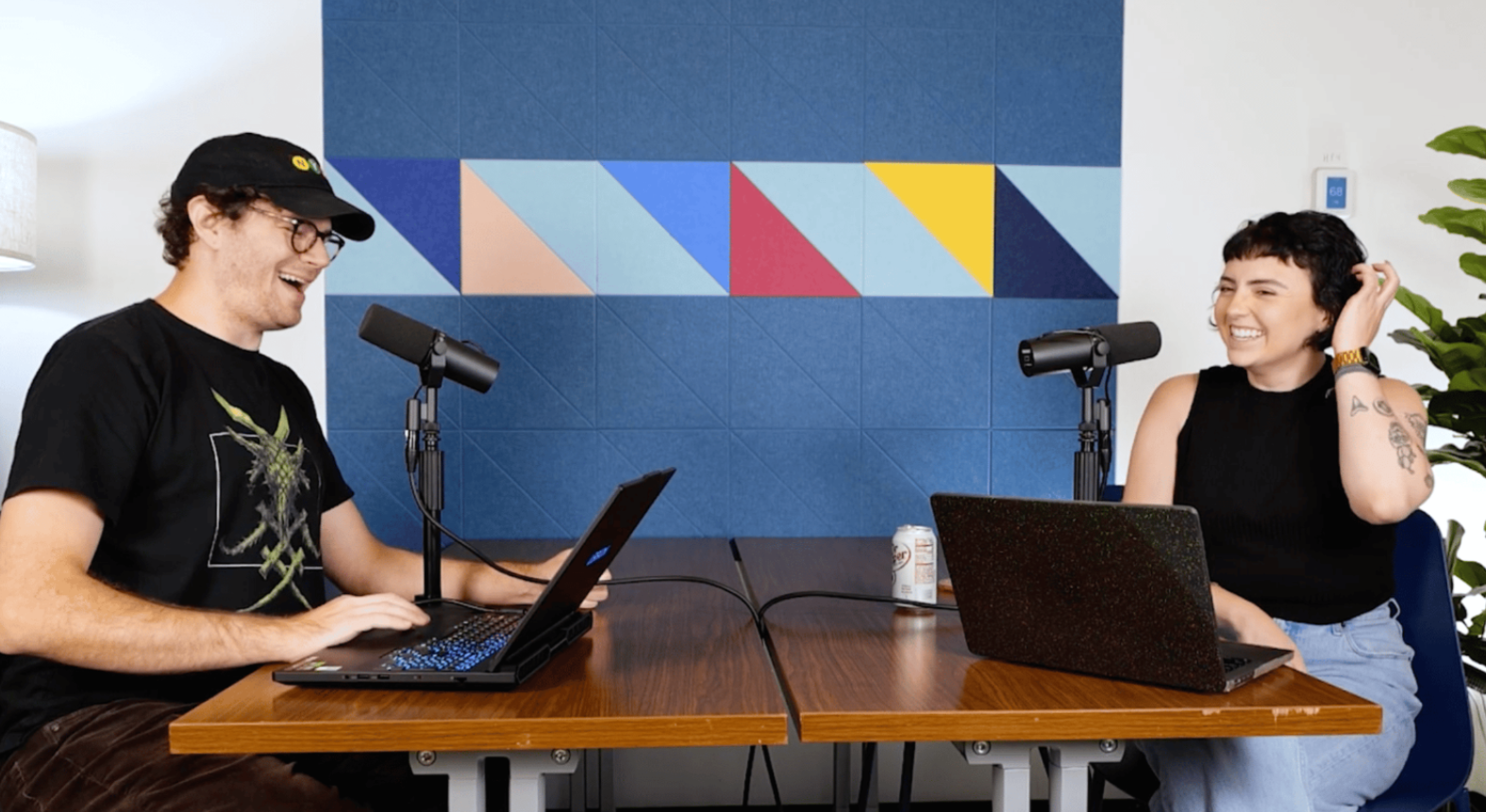 Two people sitting at a wooden table with microphones and laptops, laughing during a podcast recording, with a colorful geometric wall in the background and a potted plant on the side.